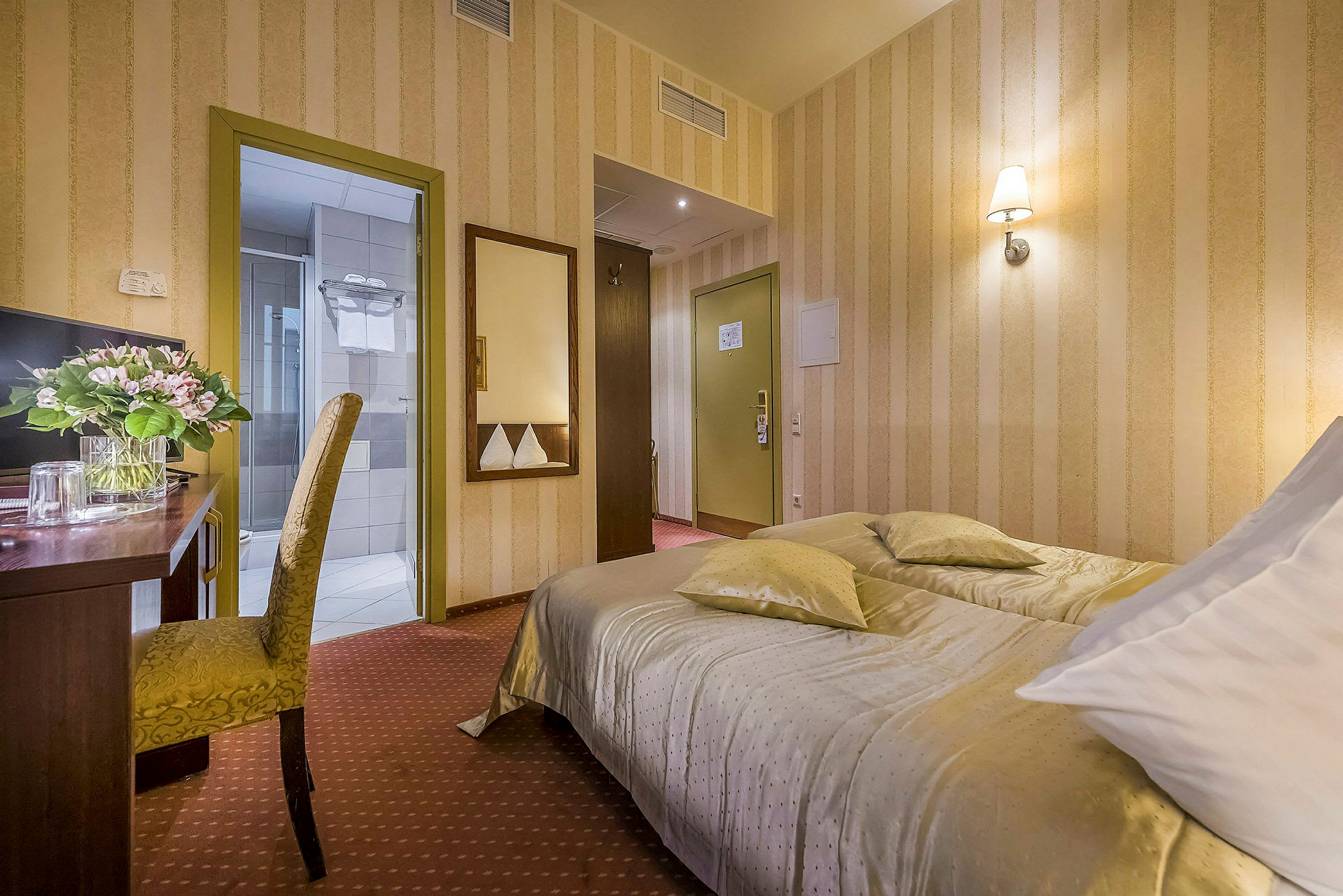 Hotellihuone, jossa pedattu sänky, kirjoituspöytä kukkamaljakolla ja tuoli sekä ovet kylpyhuoneeseen ja käytävään. (Tekoälyn luoma teksti)