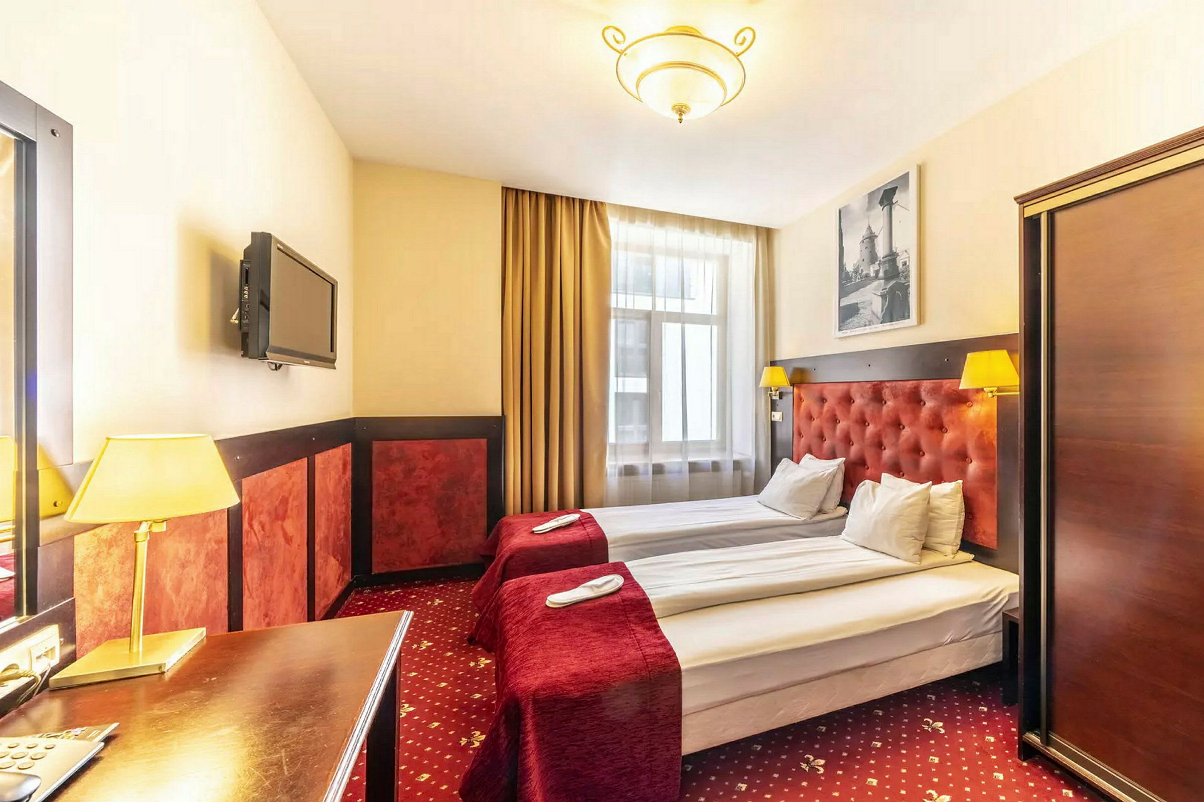 Hotellihuone, jossa kaksi yhden hengen sänkyä, punaiset päiväpeitot ja valaisimet. (Tekoälyn luoma teksti)