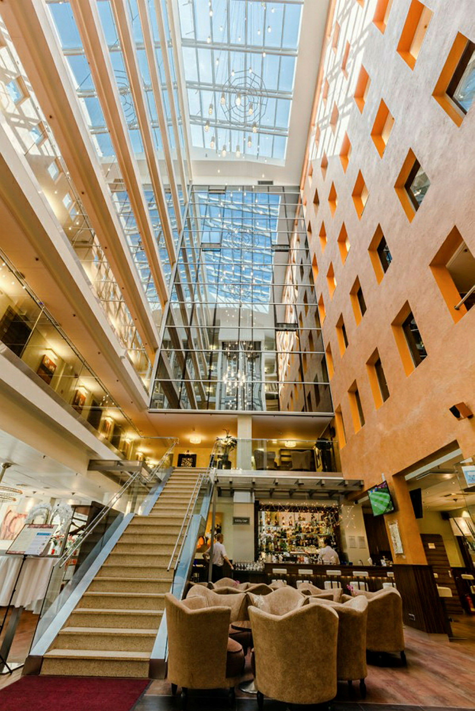 Kuva valoisasta hotelliaulasta, jossa on portaat, istuinryhmiä ja vastaanotto, luonnonvalon valaisema kattoikkunan kautta. (Tekoälyn luoma teksti)