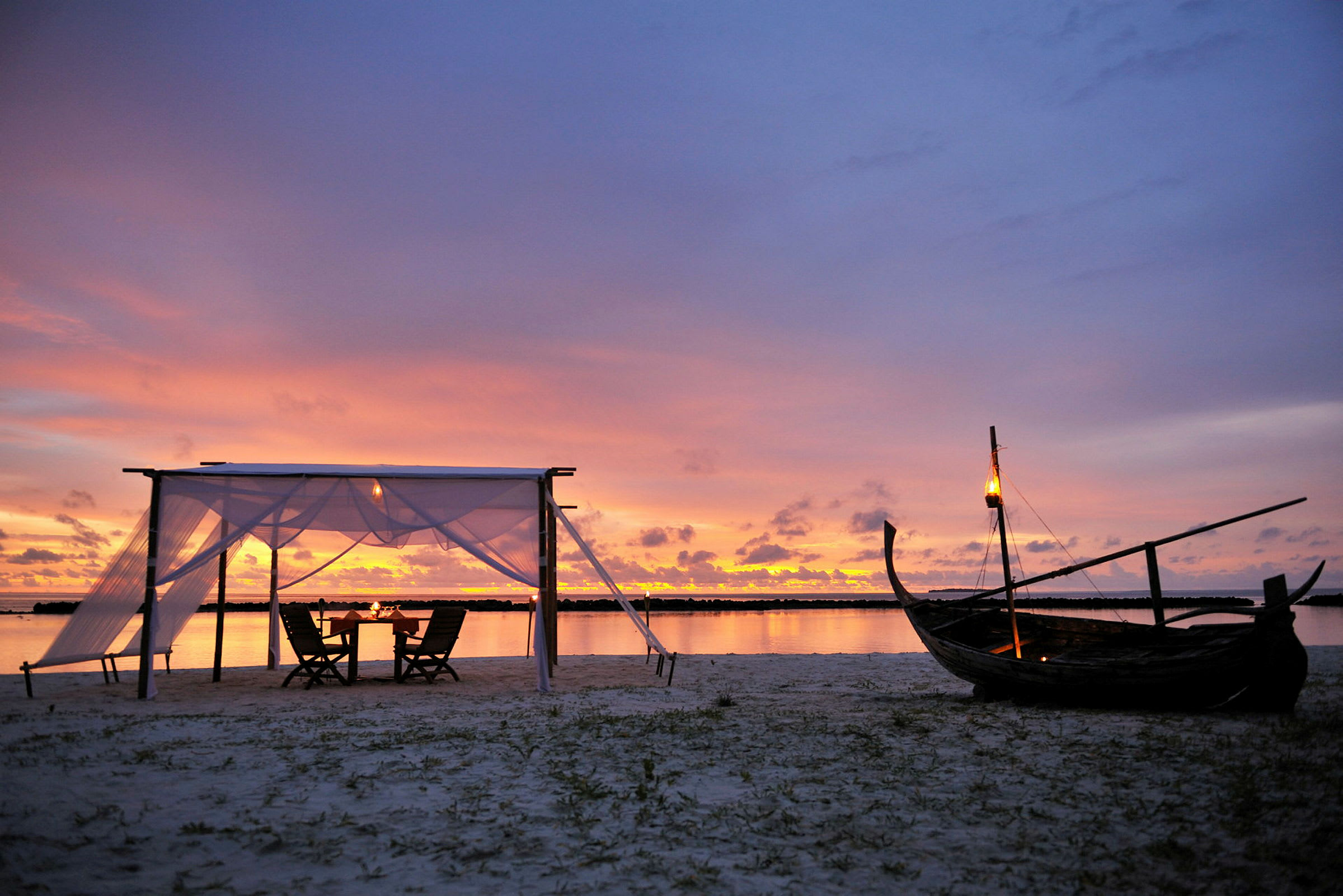 Ranta, jossa pieni katos ja tuolit auringonlaskun aikaan, puuvene ja lyhtyjä. (Tekoälyn luoma teksti)