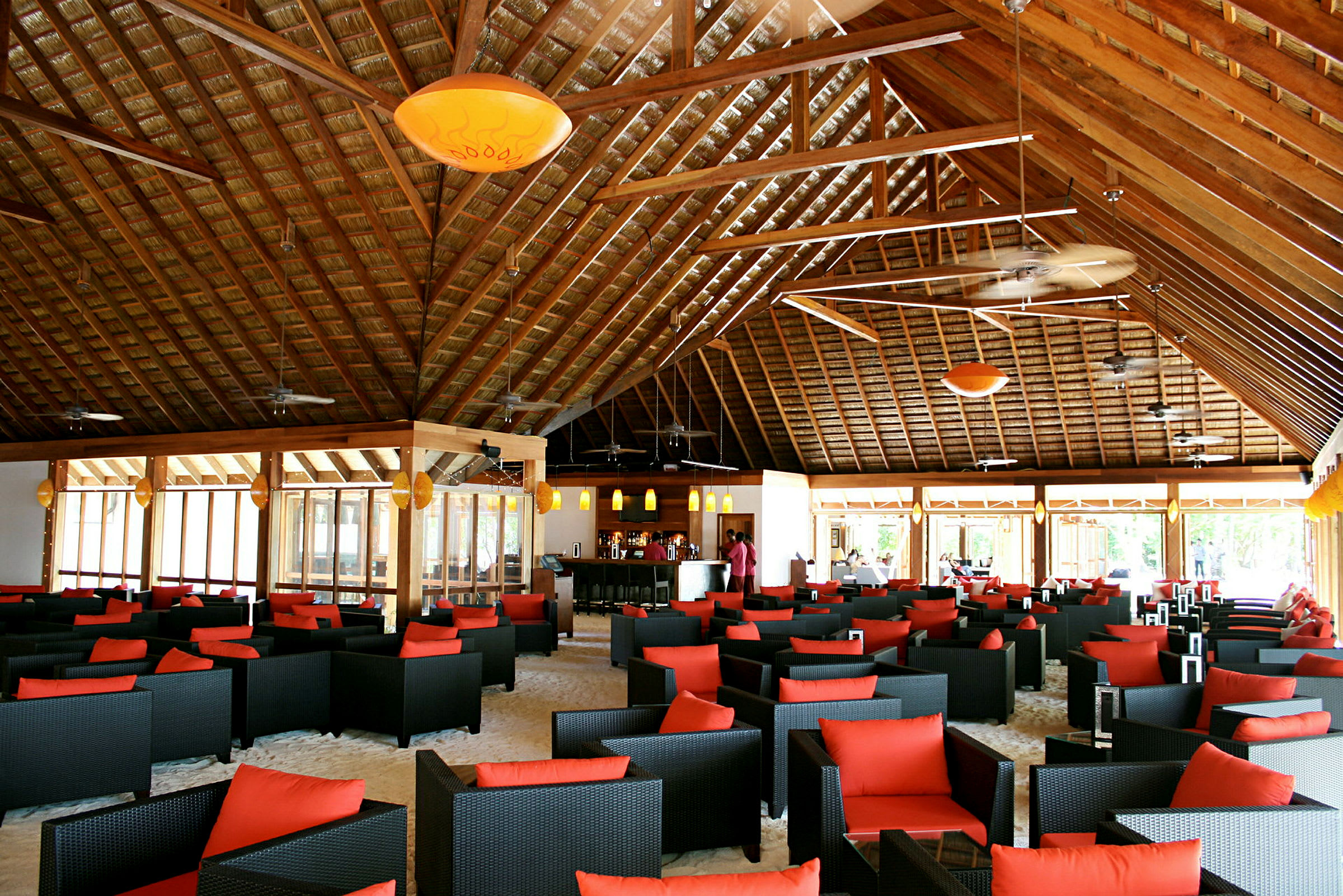 Tilava ravintola tai lounge, jossa on mustia punotyöllisiä tuoleja punaisilla tyynyillä, pöydät sijoitettu hiekkapohjalle korkean puupalkkikaton alla. (Tekoälyn luoma teksti)