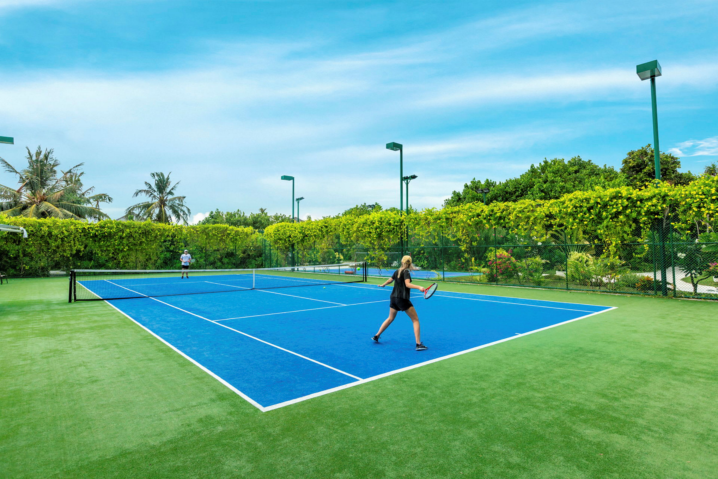 Kaksi henkilöä pelaa tennistä sinisellä tennisareenalla, jota ympäröi vihreä kasvillisuus. (Tekoälyn luoma teksti)