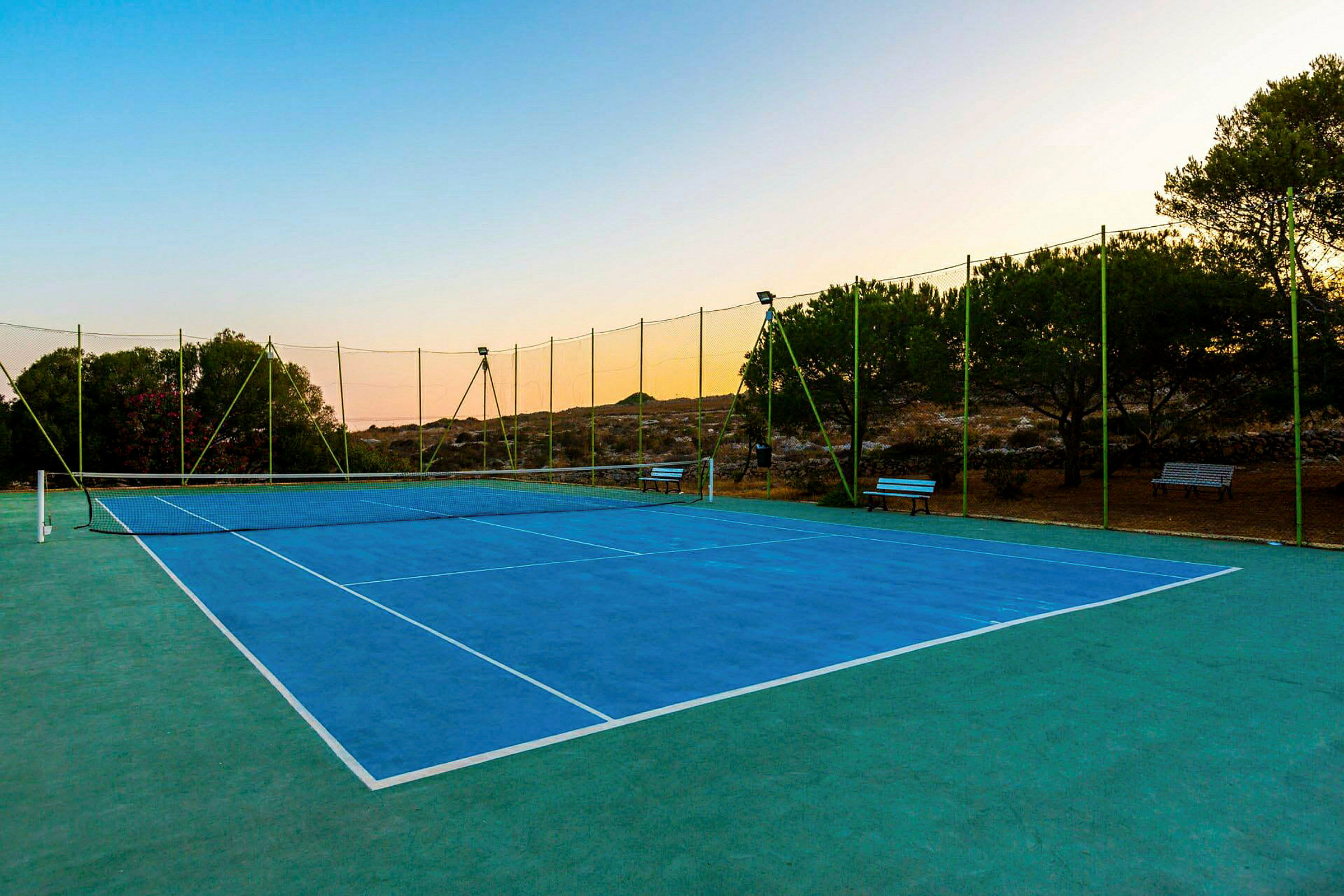 Tenniskenttä, jossa sininen pinnoite, ympäröity vihreällä verkolla ja puilla. (Tekoälyn luoma teksti)