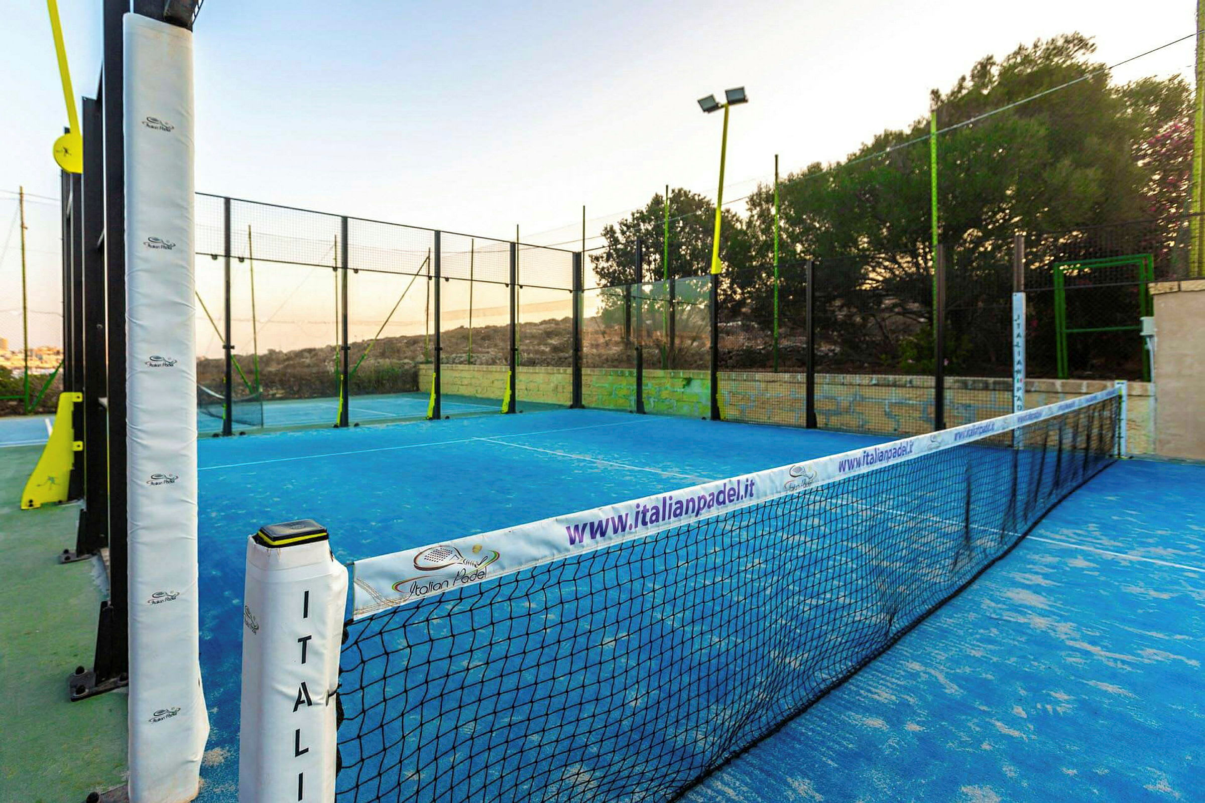 Padelkenttä, jossa on siniset merkinnät ja jota ympäröi verkko sekä aita ulkoilmassa. (Tekoälyn luoma teksti)