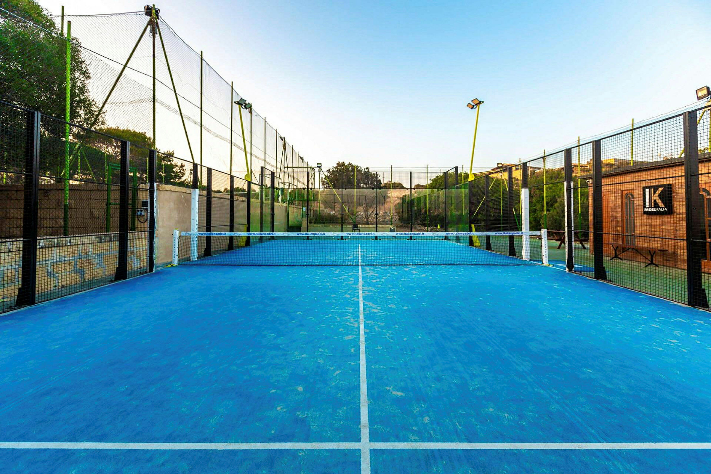 Padelkenttä sinisellä pinnalla ja verkolla keskellä, aidan ympäröimänä. (Tekoälyn luoma teksti)
