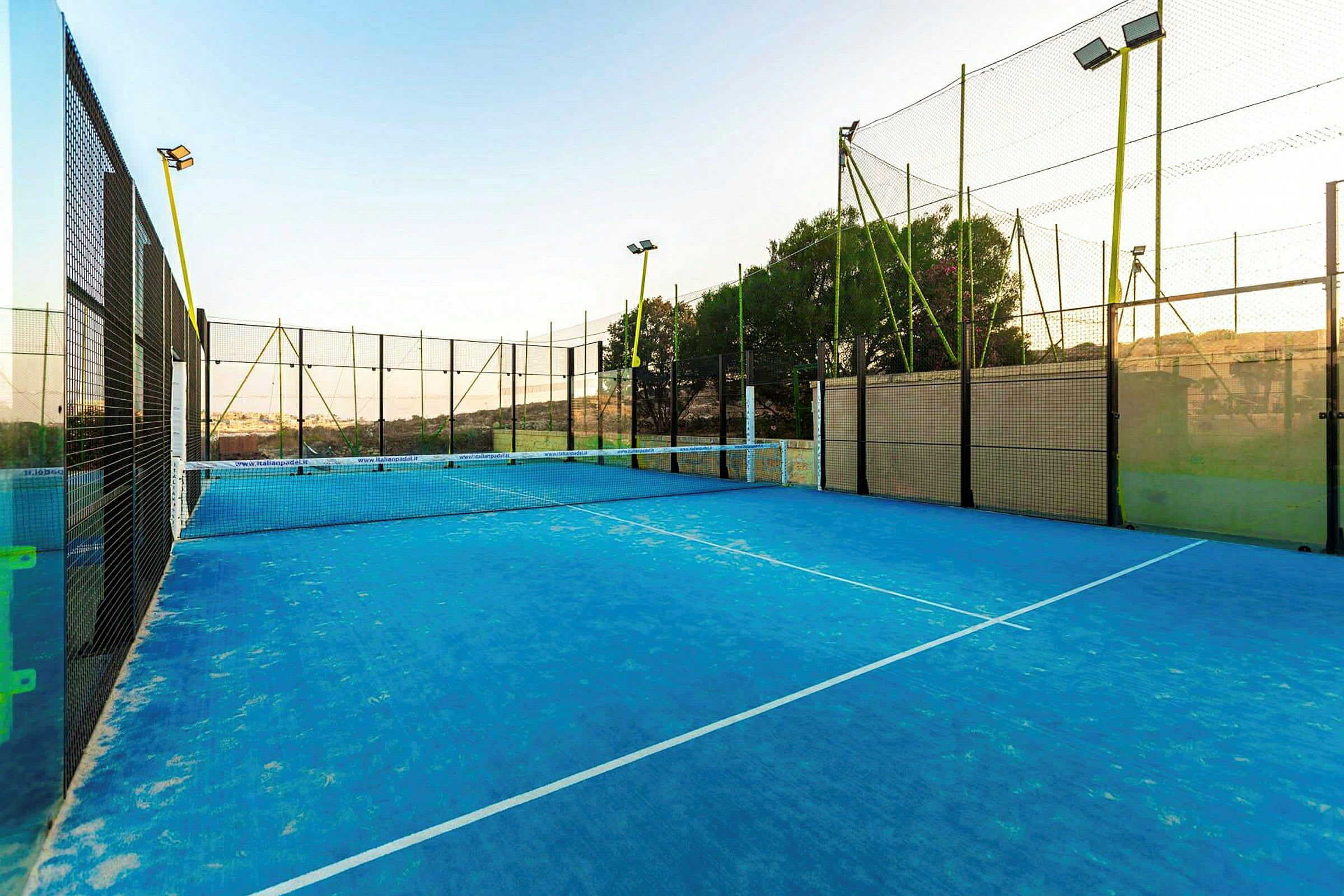 Padelkenttä, jossa on sininen alusta ja jota ympäröivät korkeat verkot, kirkkaan taivaan alla. (Tekoälyn luoma teksti)