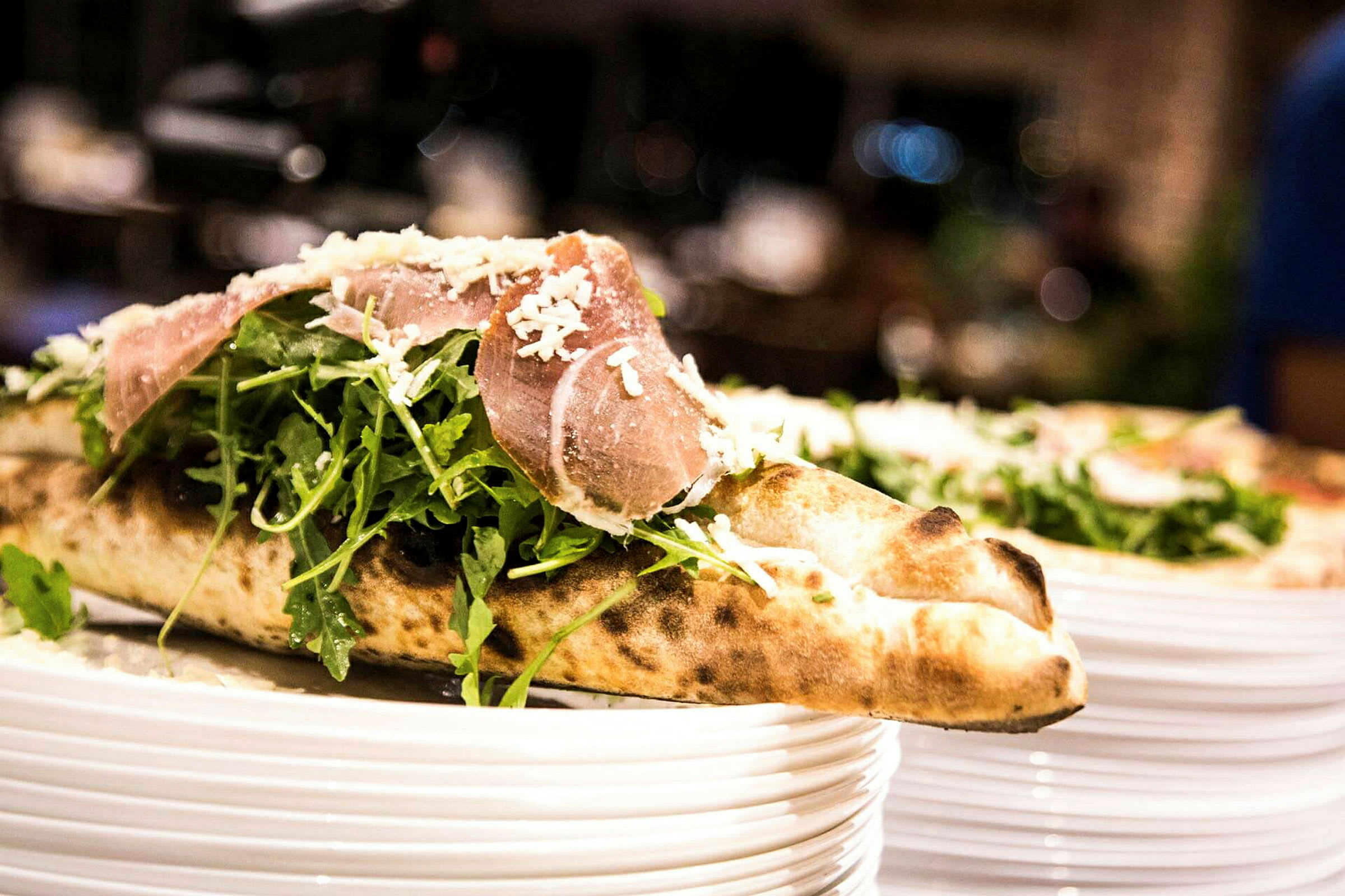 Lähikuva pizzasta, jossa on rucolasalaattia ja kinkkua, päälle raastettua juustoa, tarjoiltuna lautasella. (Tekoälyn luoma teksti)