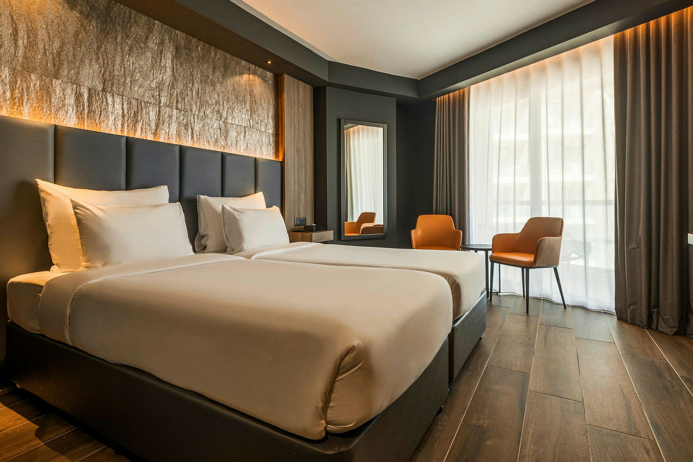 Hotellihuone, jossa on kaksi erillistä vuodetta, suuri ikkunaseinä verhoilla, kaksi oranssia tuolia ja pöytä. (Tekoälyn luoma teksti)