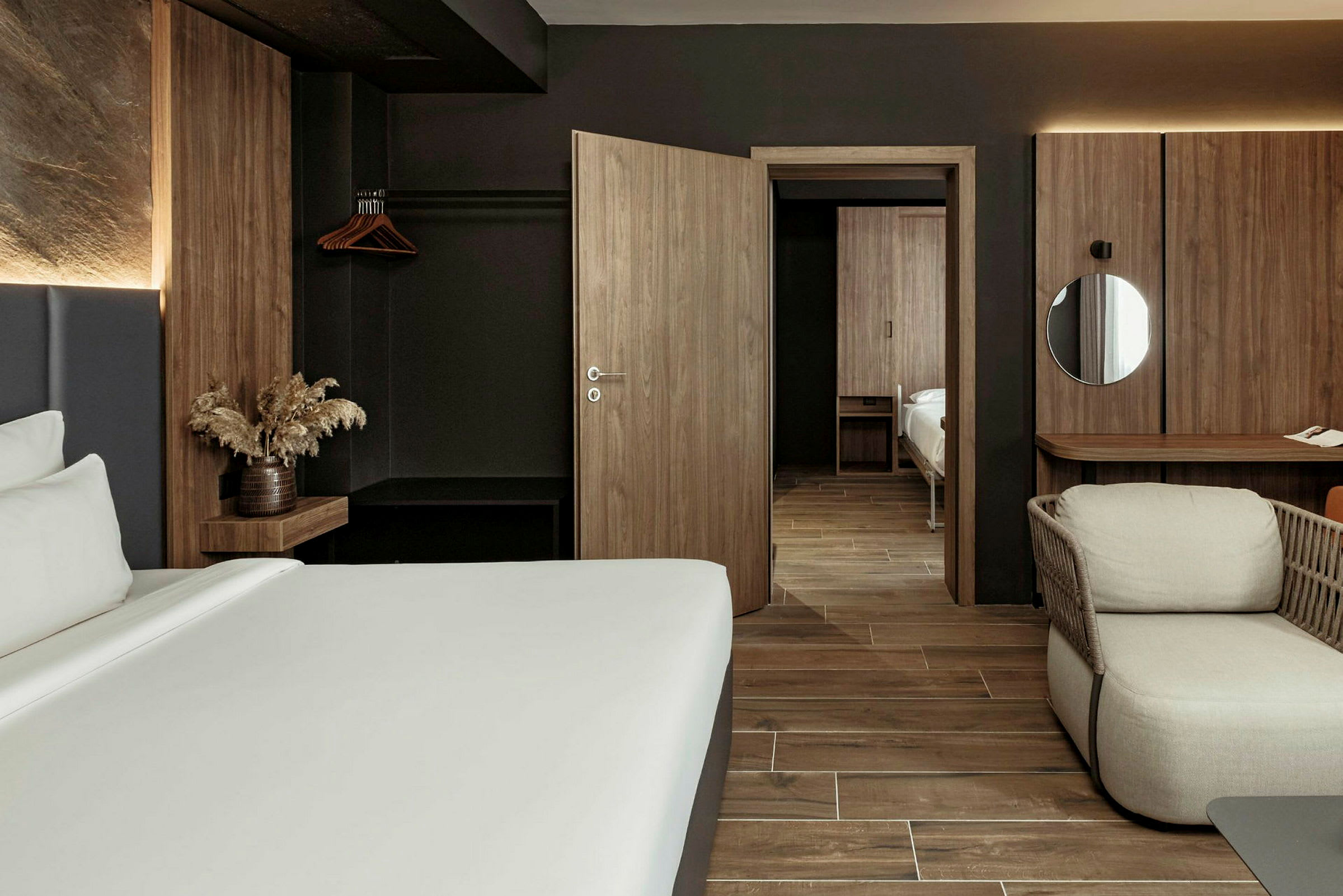Moderni hotellihuone, jossa on tummia puukalusteita ja minimalistinen sisustus. Huoneessa näkyy sänky, nojatuoli ja kirjoituspöytä. (Tekoälyn luoma teksti)