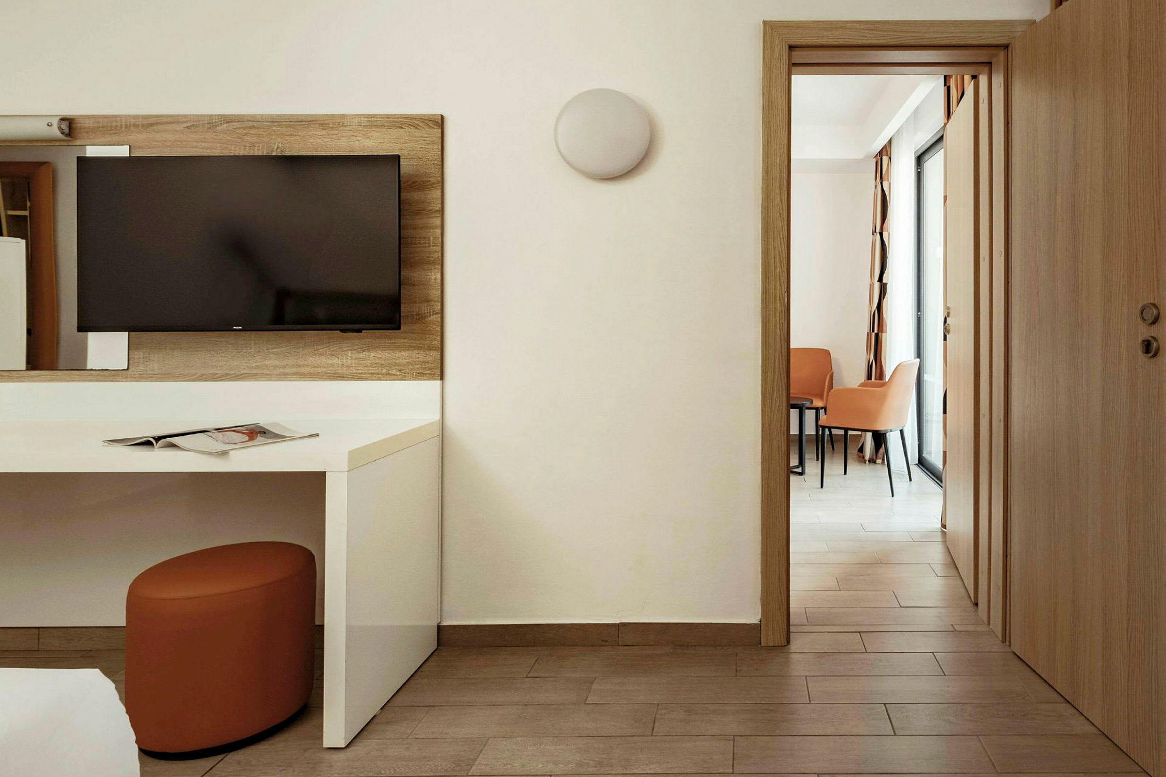 Hotellihuone, jossa seinällä on televisio valkoisen pöydän yläpuolella. Ovi avautuu alueelle, jossa on kaksi tuolia ja pöytä. (Tekoälyn luoma teksti)