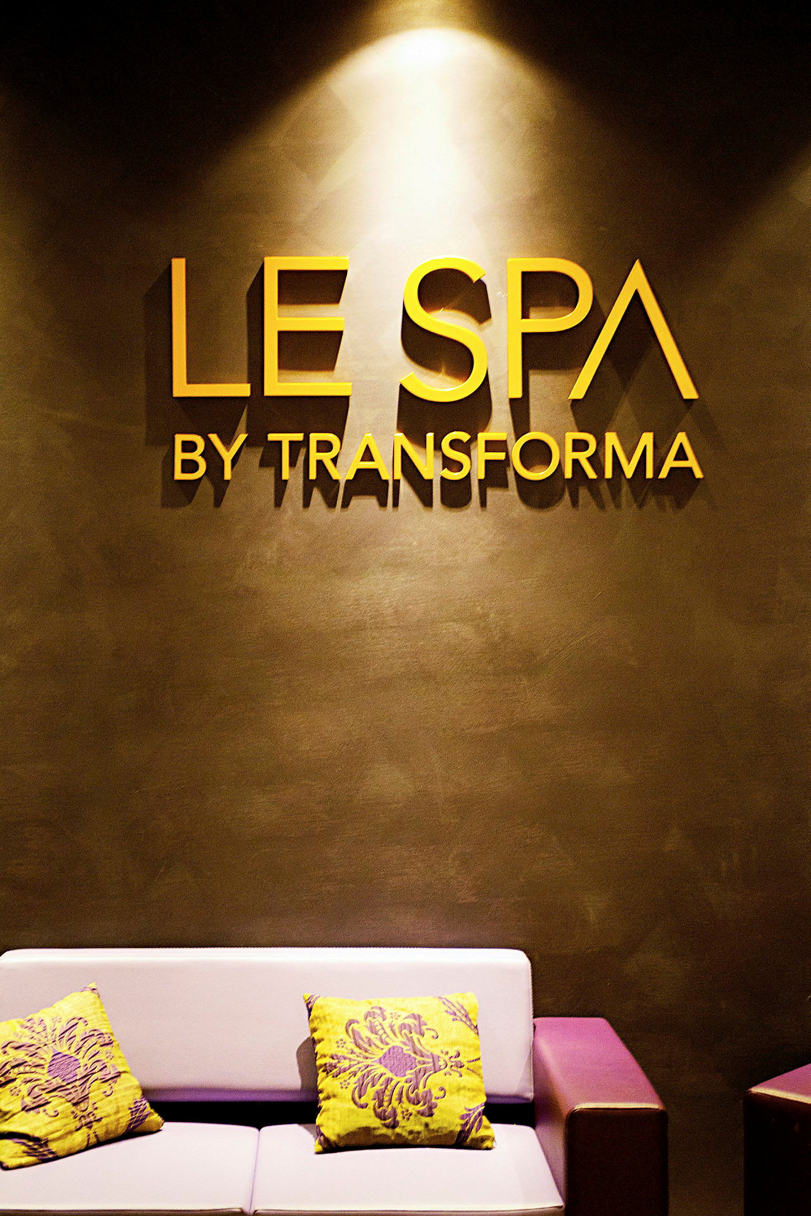 Kuva kylpylän sisäänkäynnistä, jossa seinällä lukee "Le Spa by Transforma". Näkyvissä on sohva, jossa on värikkäitä koristetyynyjä. (Tekoälyn luoma teksti)