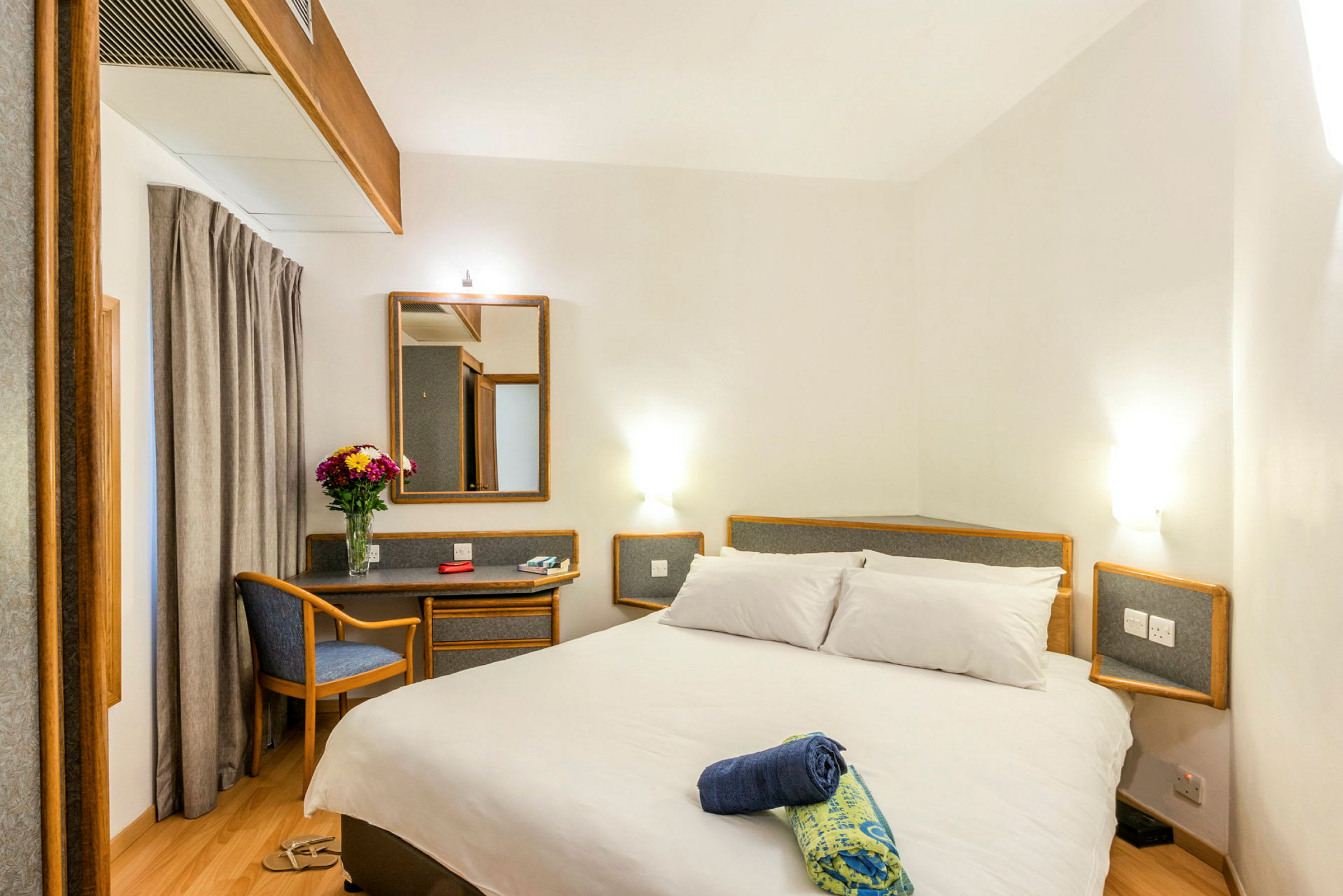 Hotellihuone, jossa on parisänky, peili, pieni työpöytä ja tuoli. Työpöydällä on maljakko kukkia. (Tekoälyn luoma teksti)