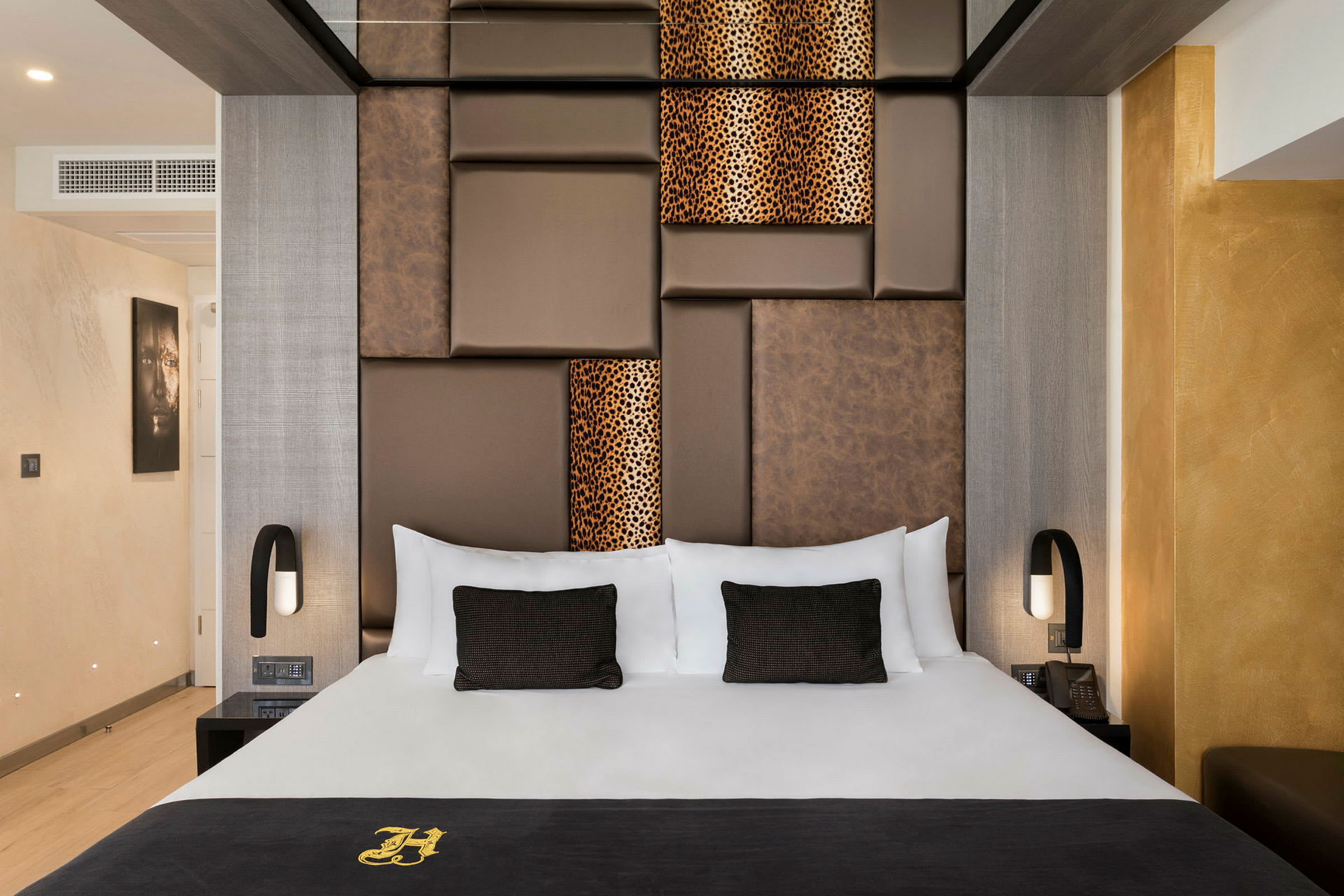 Tyylikäs hotellihuone, jossa on suuri sänky ja ruskeita sekä kuvioituja paneeleja seinällä takana. Huone on modernisti sisustettu lämpimillä väreillä. (Tekoälyn luoma teksti)