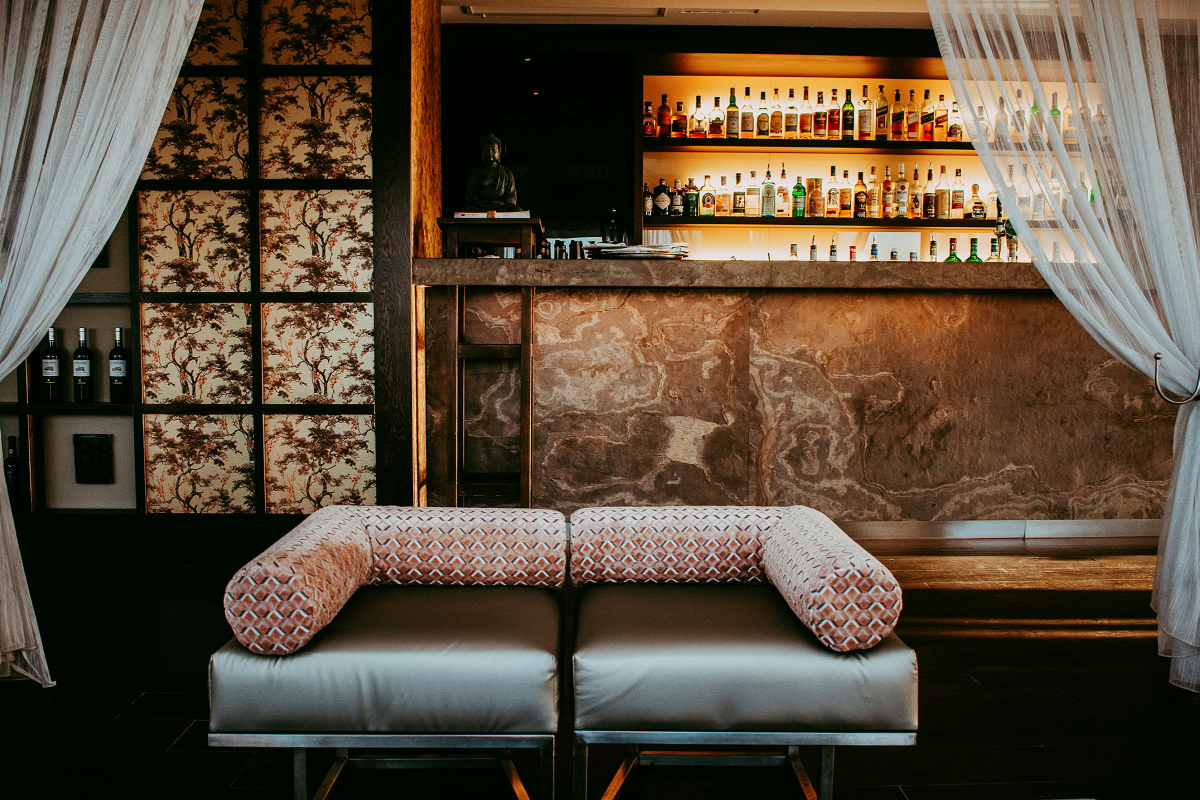 Lounge-kaluste baaritiskin edessä, jonka hyllyt ovat valaistuja ja täynnä pulloja ja laseja. Seinät on koristeltu paneeleilla, ja sivuilla roikkuu verhoja. (Tekoälyn luoma teksti)