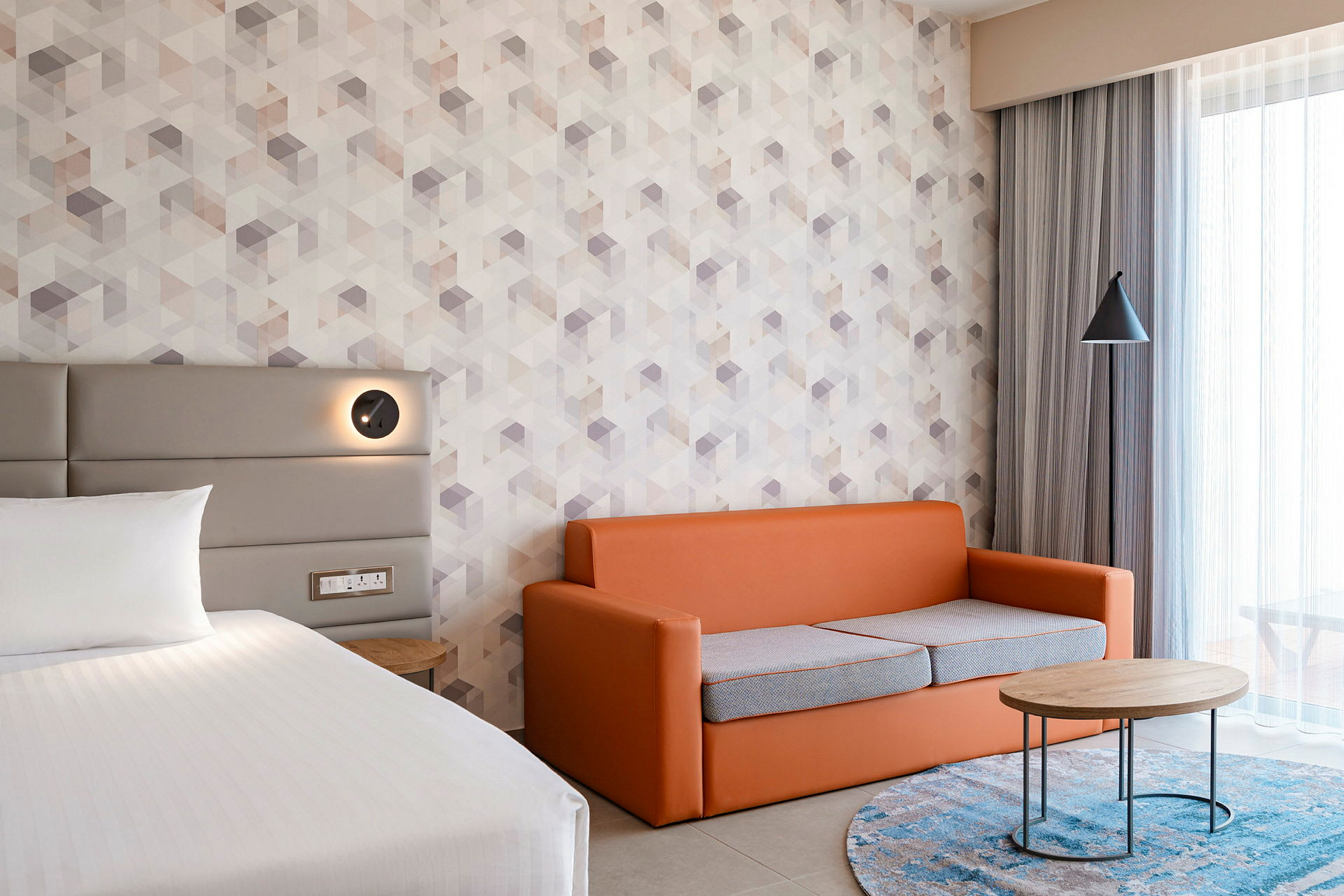 Hotellihuone, jossa on sänky, oranssi sohva ja pyöreä sohvapöytä. (Tekoälyn luoma teksti)
