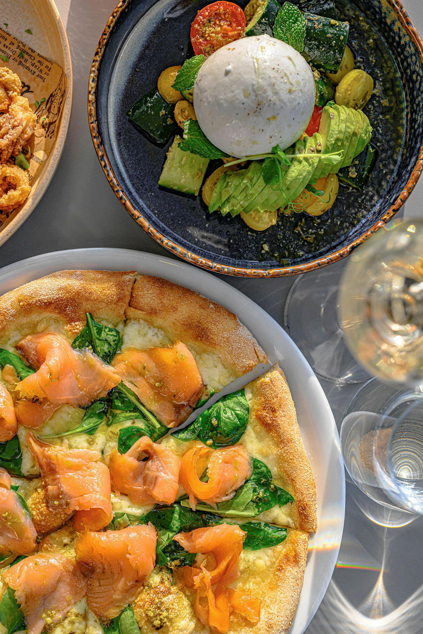 Lautanen, jossa on burrataa, vihanneksia ja avokadoa, sekä pizzaa lohella ja pinaatilla. (Tekoälyn luoma teksti)