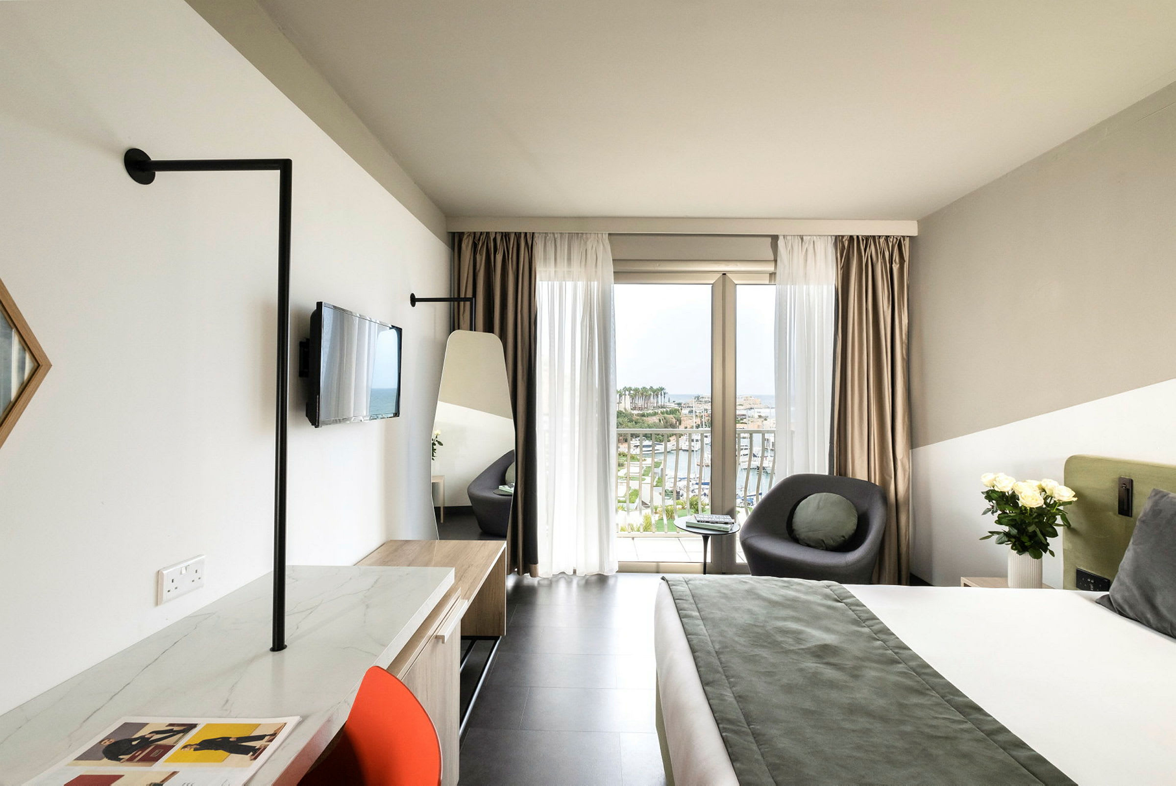 Moderni hotellihuone, jossa suuri sänky, istuinryhmä ikkunan vieressä ja näkymä satamaan sekä työpiste tuolilla. (Tekoälyn luoma teksti)