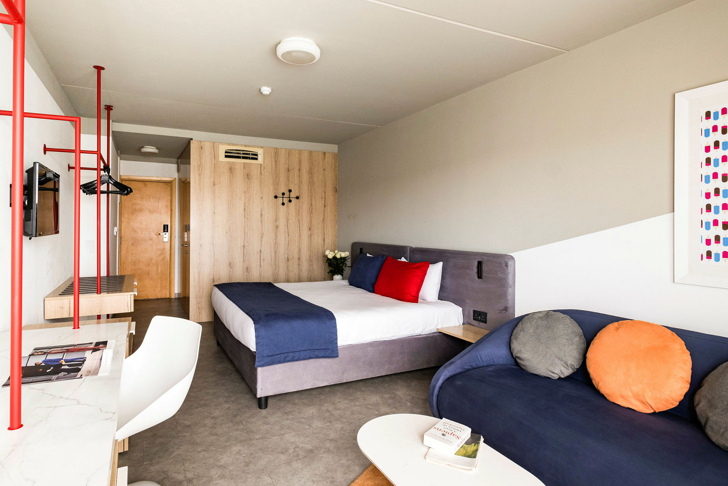 Hotellihuone, jossa on sänky sinisellä peitolla ja punaisilla ja valkoisilla tyynyillä, sininen sohva värikkäillä tyynyillä sekä puinen pöytä ja vaatekaappi. (Tekoälyn luoma teksti)