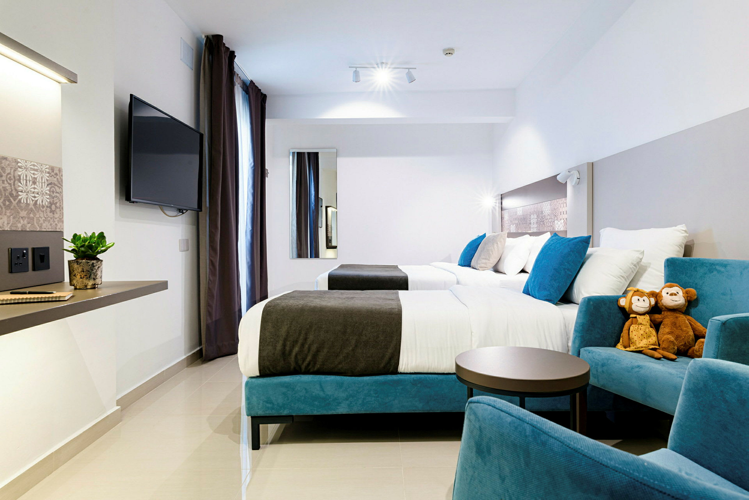 Moderni hotellihuone, jossa on kaksi sänkyä, istumapaikkoja, seinään kiinnitetty televisio ja kaksi pehmolelua tuolilla. (Tekoälyn luoma teksti)
