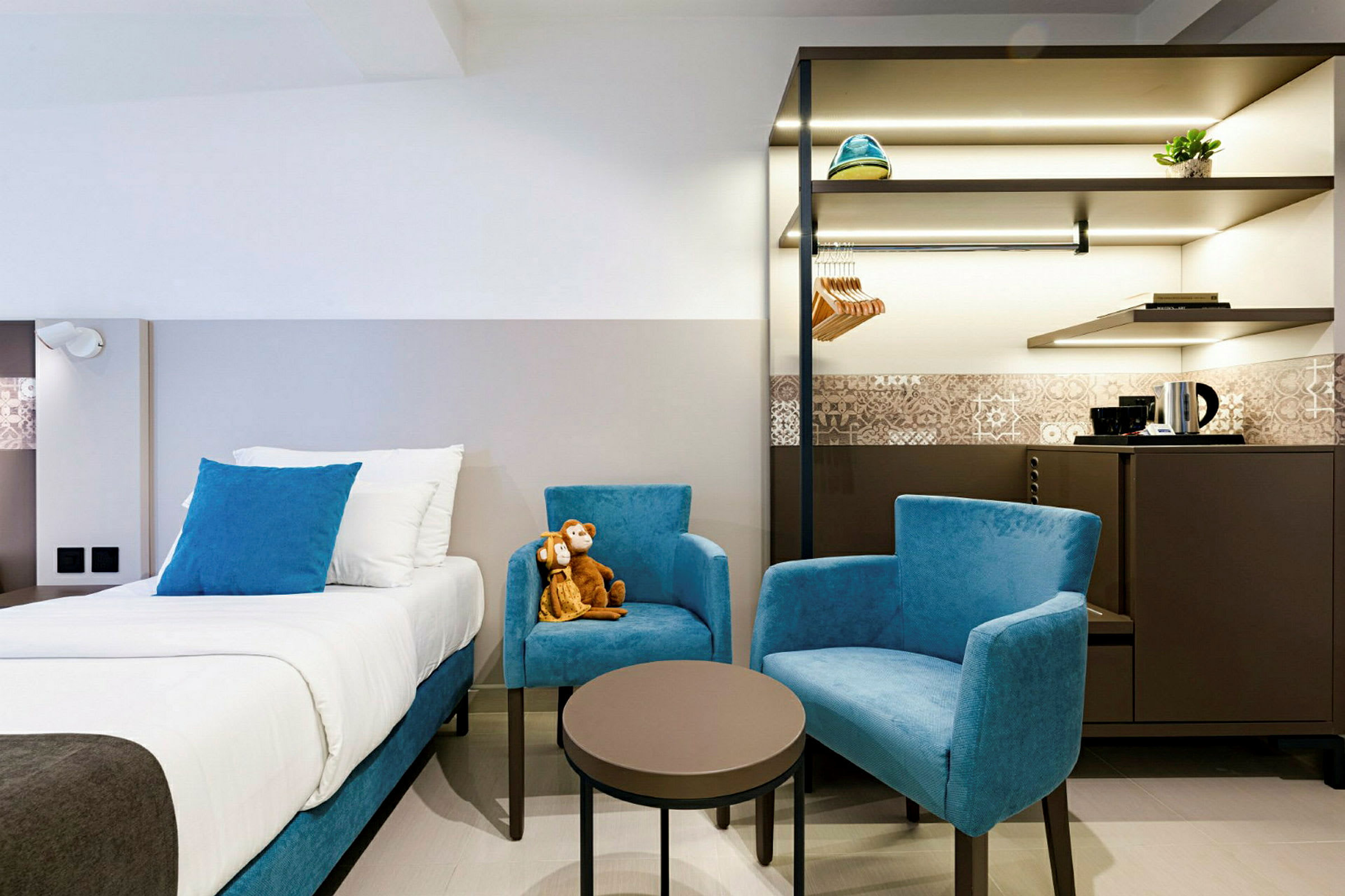 Hotellihuone, jossa on sänky, kaksi sinistä nojatuolia ja pöytä sekä kaappi ja vedenkeitin taustalla. (Tekoälyn luoma teksti)