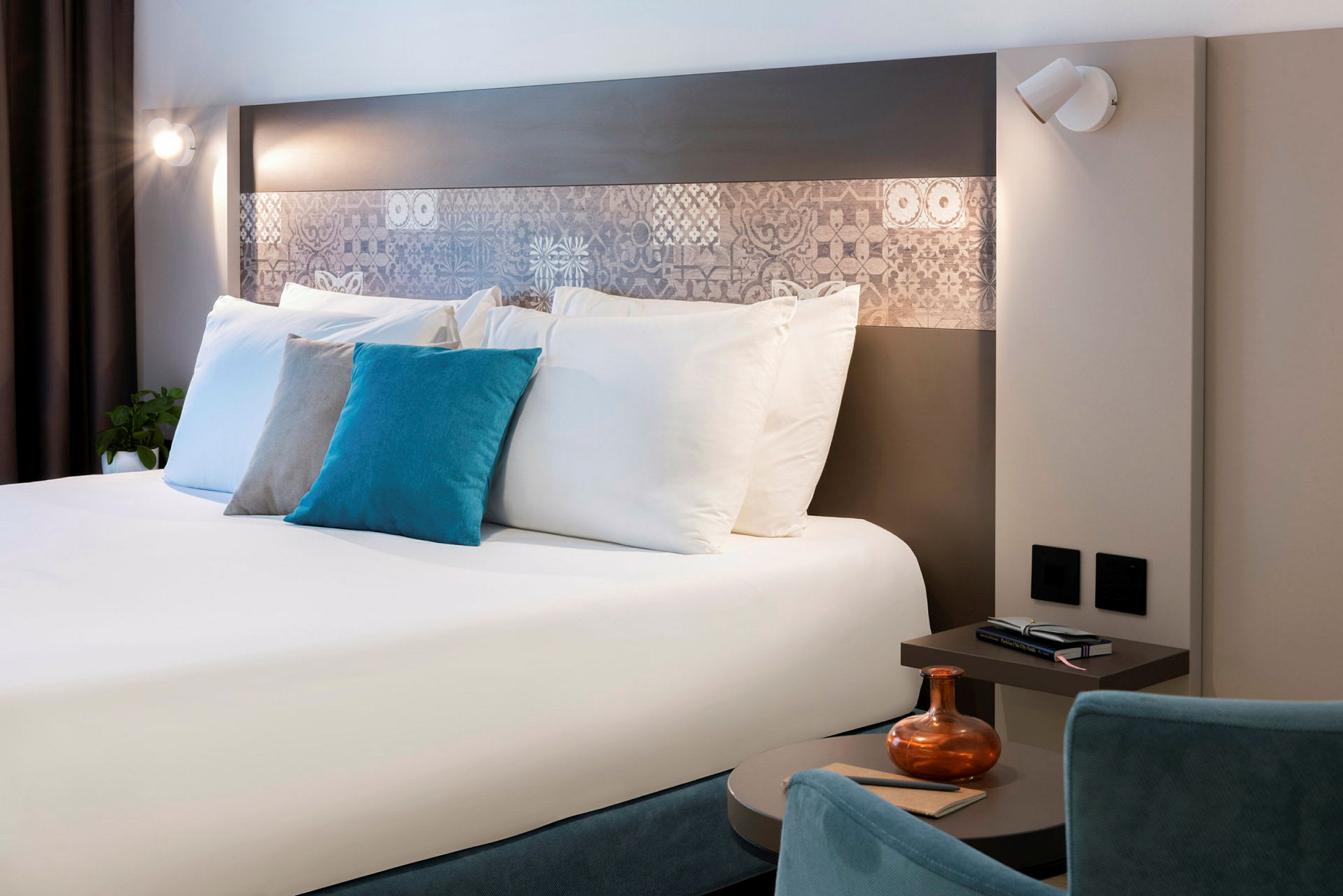Moderni hotellihuone, jossa on parivuode, koristeelliset tyynyt, yöpöytä ja valaistus. (Tekoälyn luoma teksti)