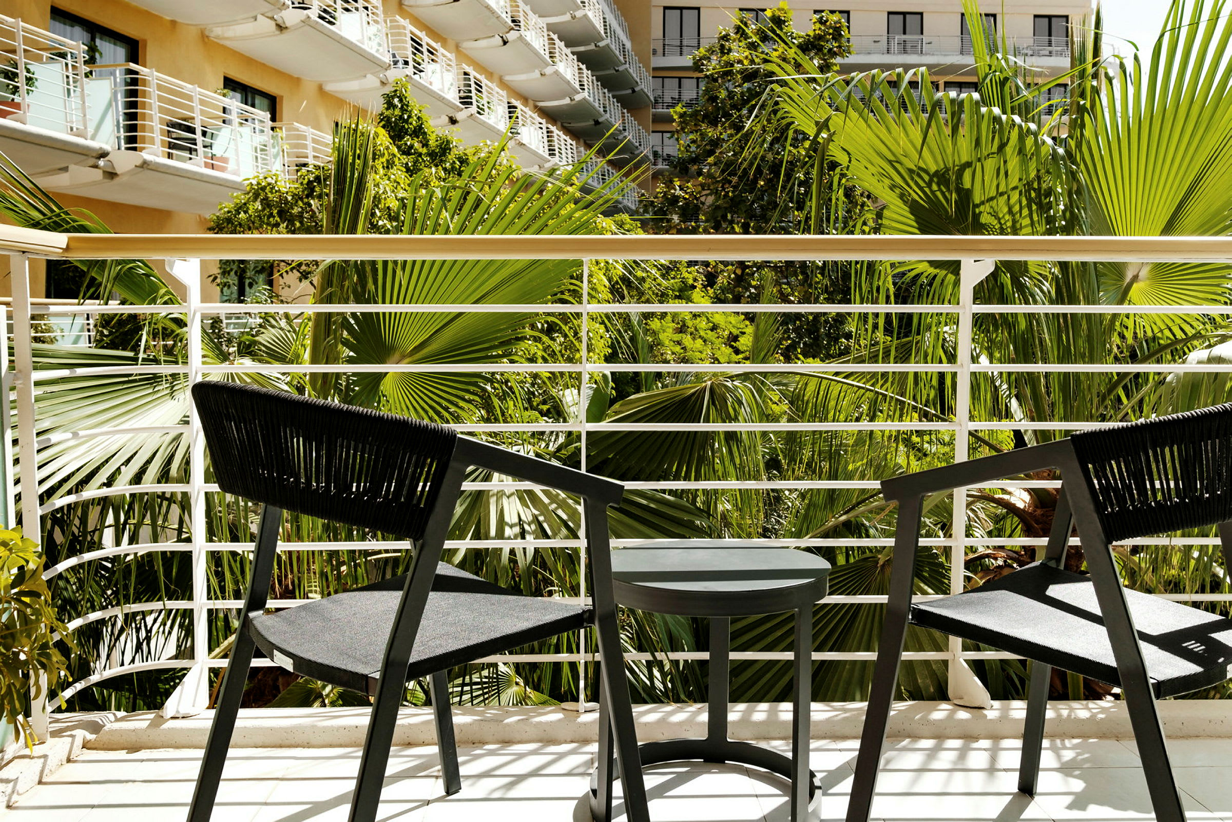 Kaksi mustaa tuolia ja pöytä parvekkeella, näkymänä vihreitä palmuja. (Tekoälyn luoma teksti)