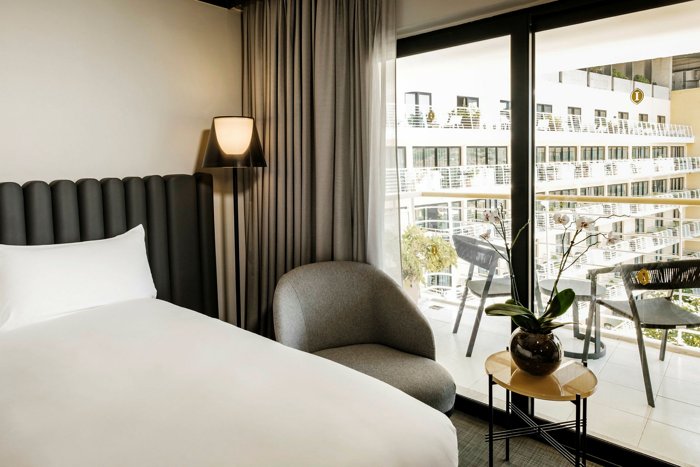 Moderni hotellihuone, jossa sänky, tuoli ja pieni pöytä kasvin kanssa, suuren ikkunan vieressä näkyvillä parvekkeilla. (Tekoälyn luoma teksti)