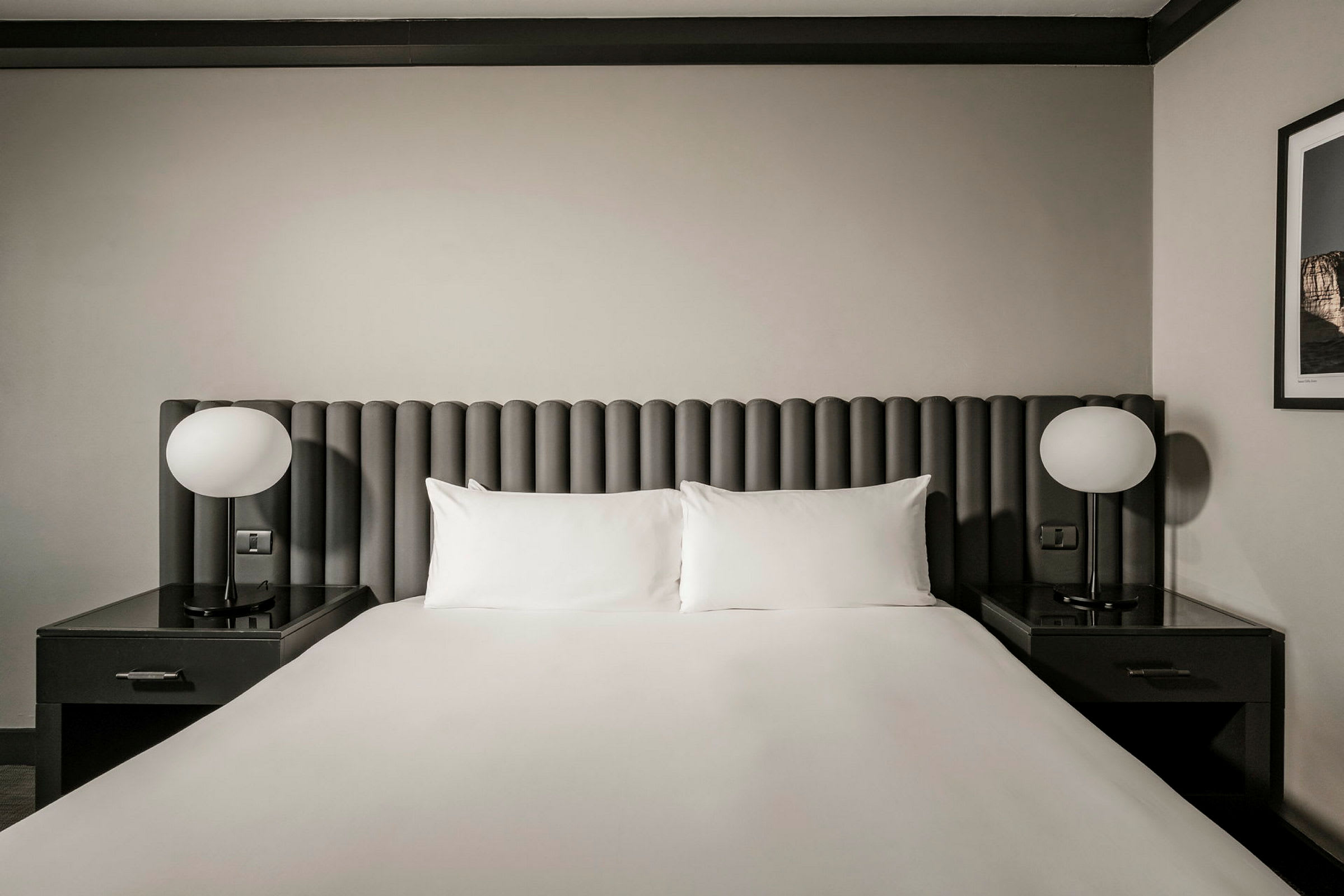 Hotellihuone, jossa on parisänky. Sängyssä on harmaa pääty, valkoiset tyynyt ja lakanat. Molemmilla puolilla on yöpöydät pyöreine lampuineen. (Tekoälyn luoma teksti)