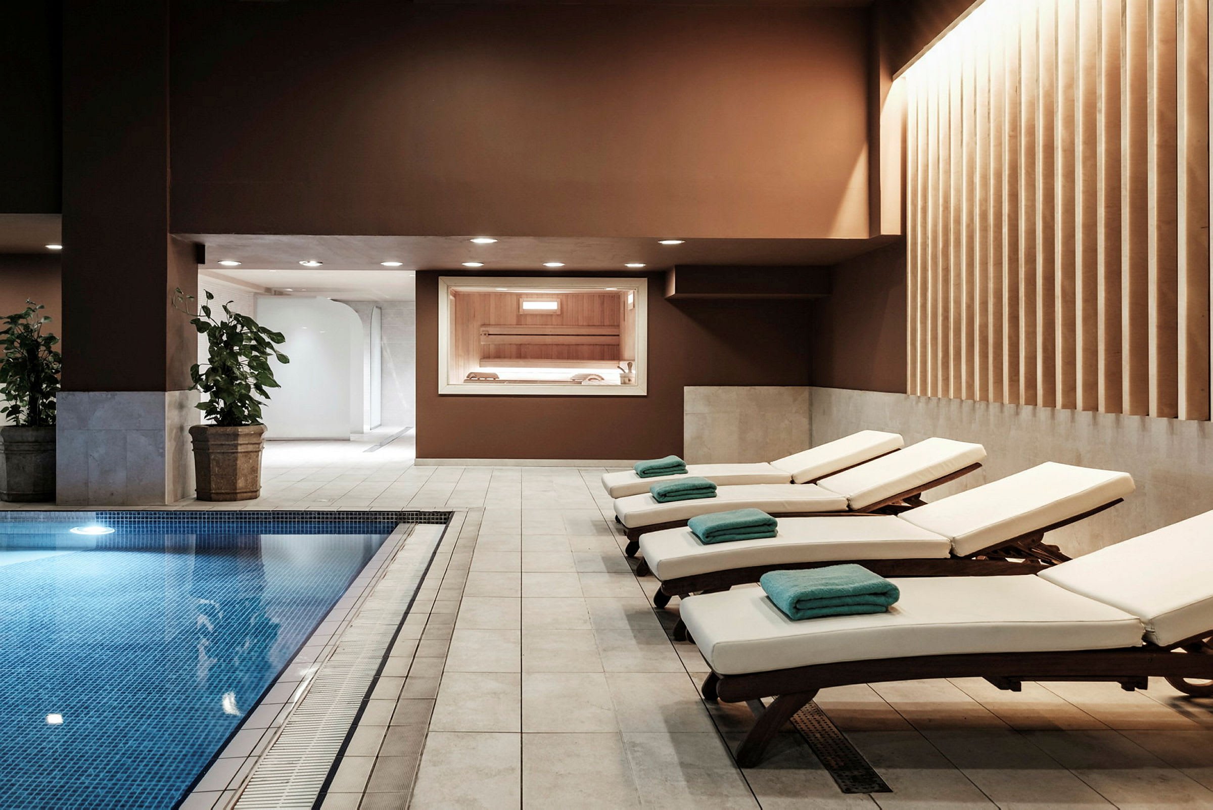 Sisäkuva spa-alueesta, jossa on uima-allas, aurinkotuoleja pyyhkeineen ja sauna näkyy ikkunan läpi. (Tekoälyn luoma teksti)