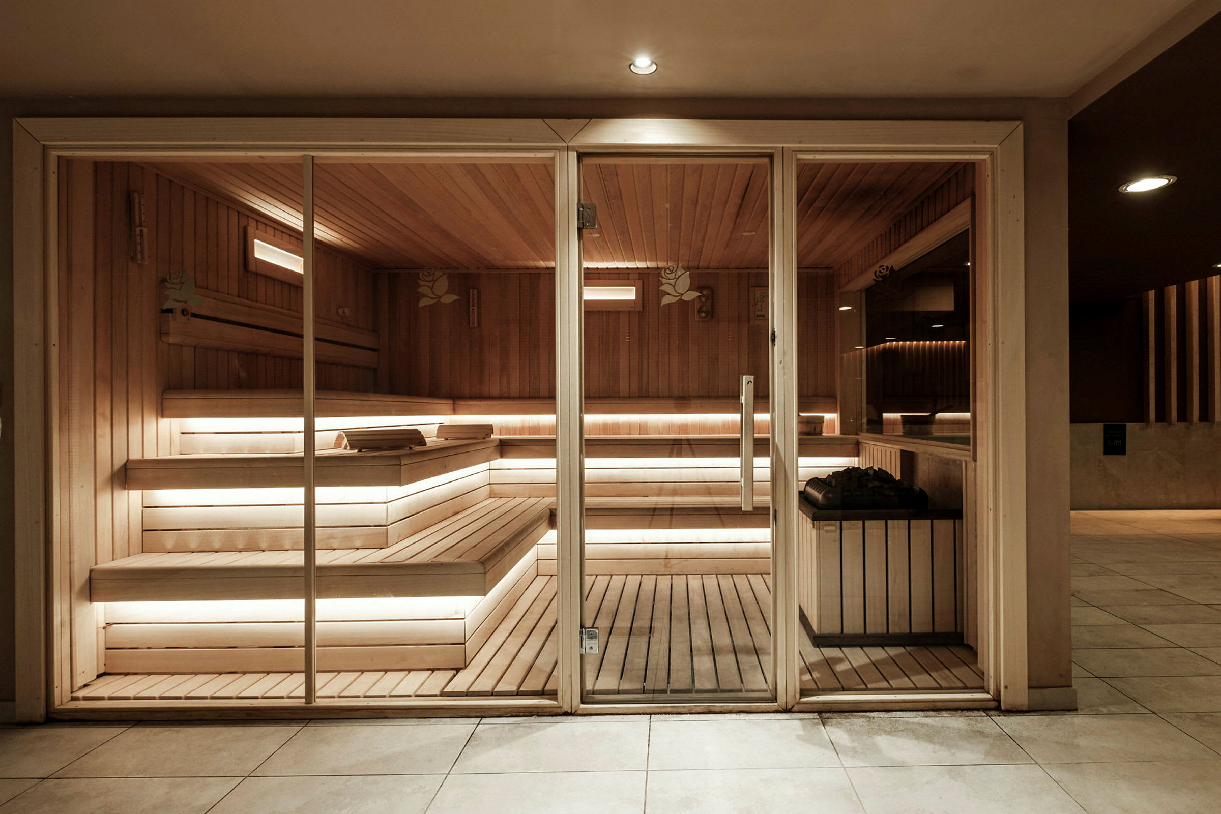 Sauna, jossa on puukalusteet, lasiovi ja penkit sekä valaistus. (Tekoälyn luoma teksti)