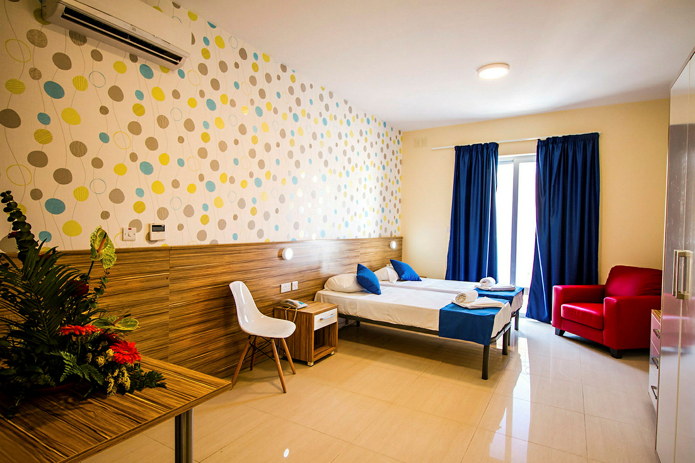 Hotellihuone, jossa kaksi yhden hengen vuodetta, siniset vuodevaatteet, pöytä kukilla, valkoinen tuoli ja punainen nojatuoli. Seinää koristaa värikäs kuvio. (Tekoälyn luoma teksti)