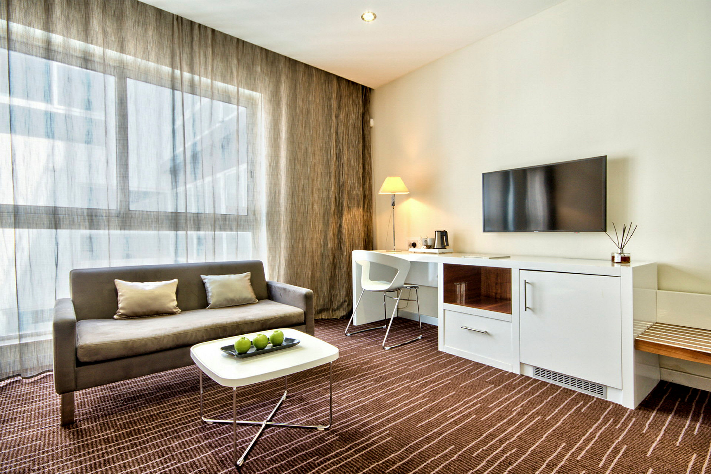 Moderni hotellihuone, jossa on sohva, sohvapöytä vihreillä omenilla, työtila tuolilla ja lampulla sekä seinälle asennettu televisio. (Tekoälyn luoma teksti)