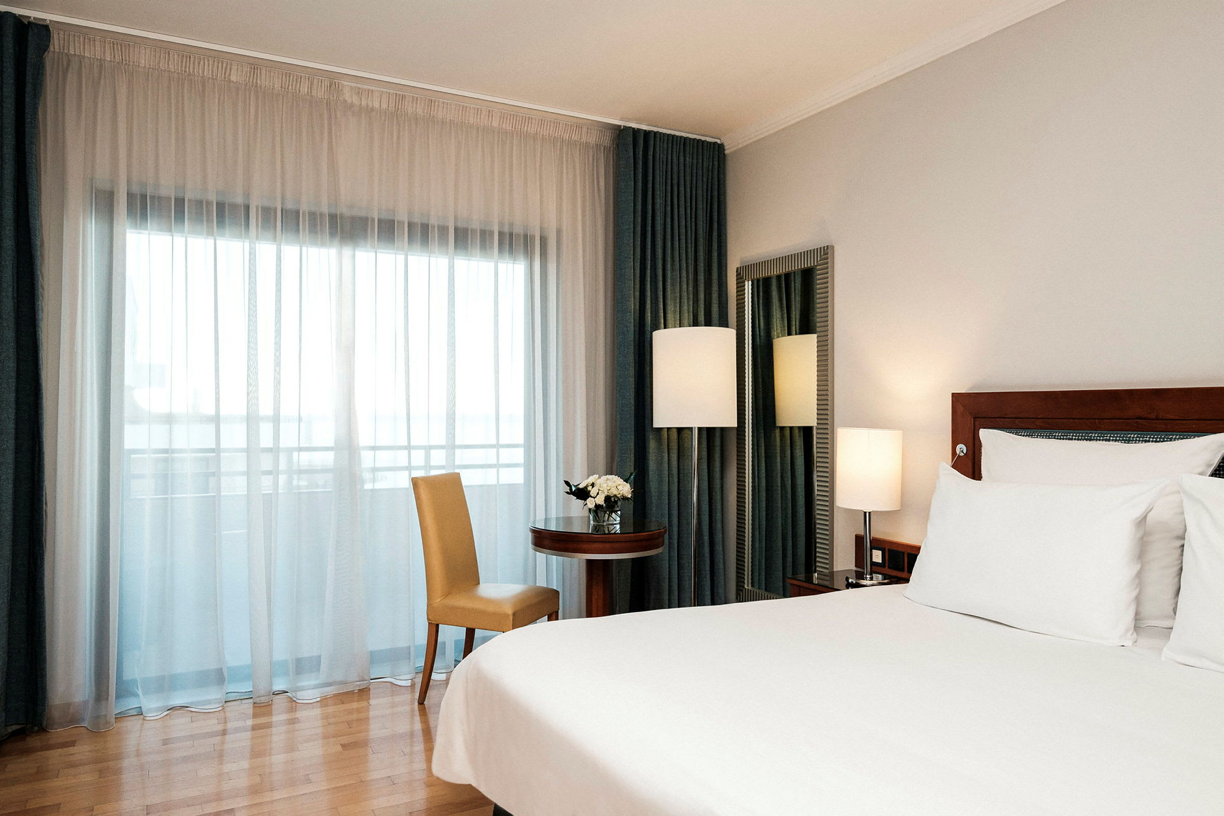 Hotellihuone, jossa on iso sänky, pöytä ja tuoli sekä ikkuna valkoisilla verhoilla. (Tekoälyn luoma teksti)