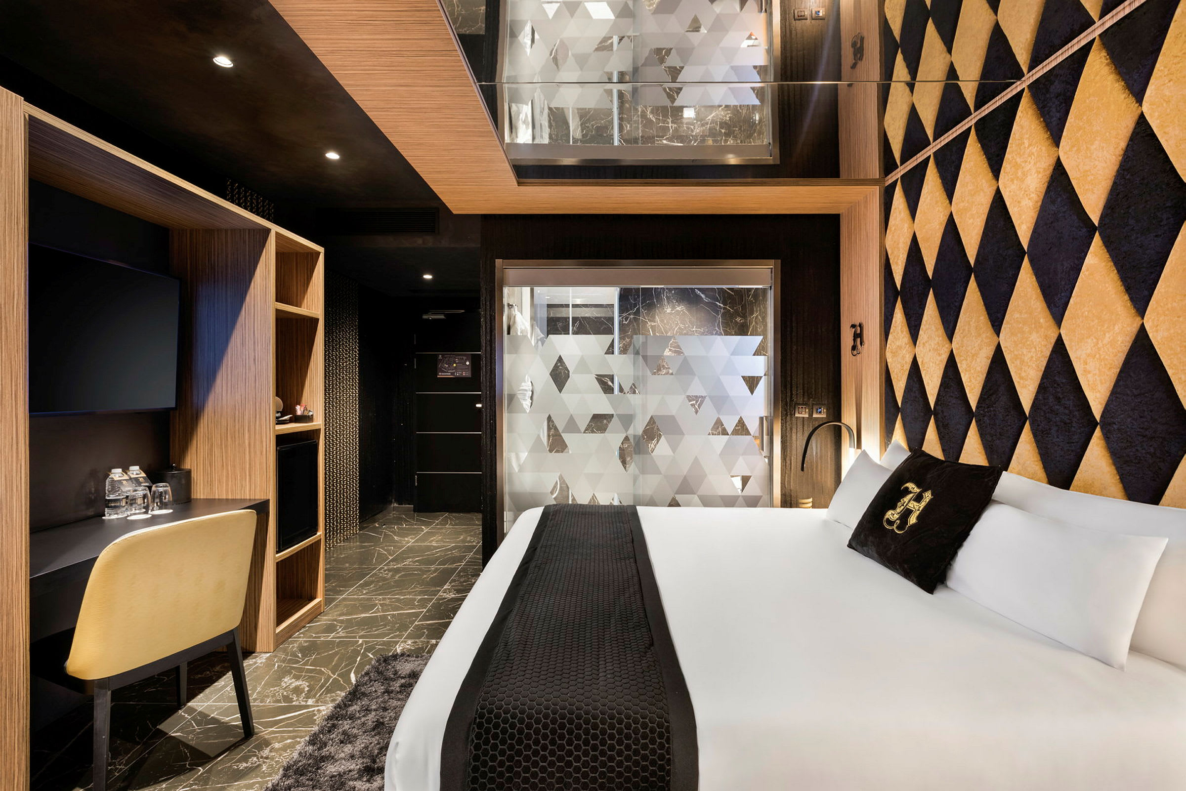 Modernisti sisustettu hotellihuone. Huoneessa on suuri sänky, geometrisiä koristeellisia seinäpintoja sekä kirjoituspöytä ja tuoli. Huurrettu lasiseinä erottaa kylpyhuoneen makuualueesta. (Tekoälyn luoma teksti)