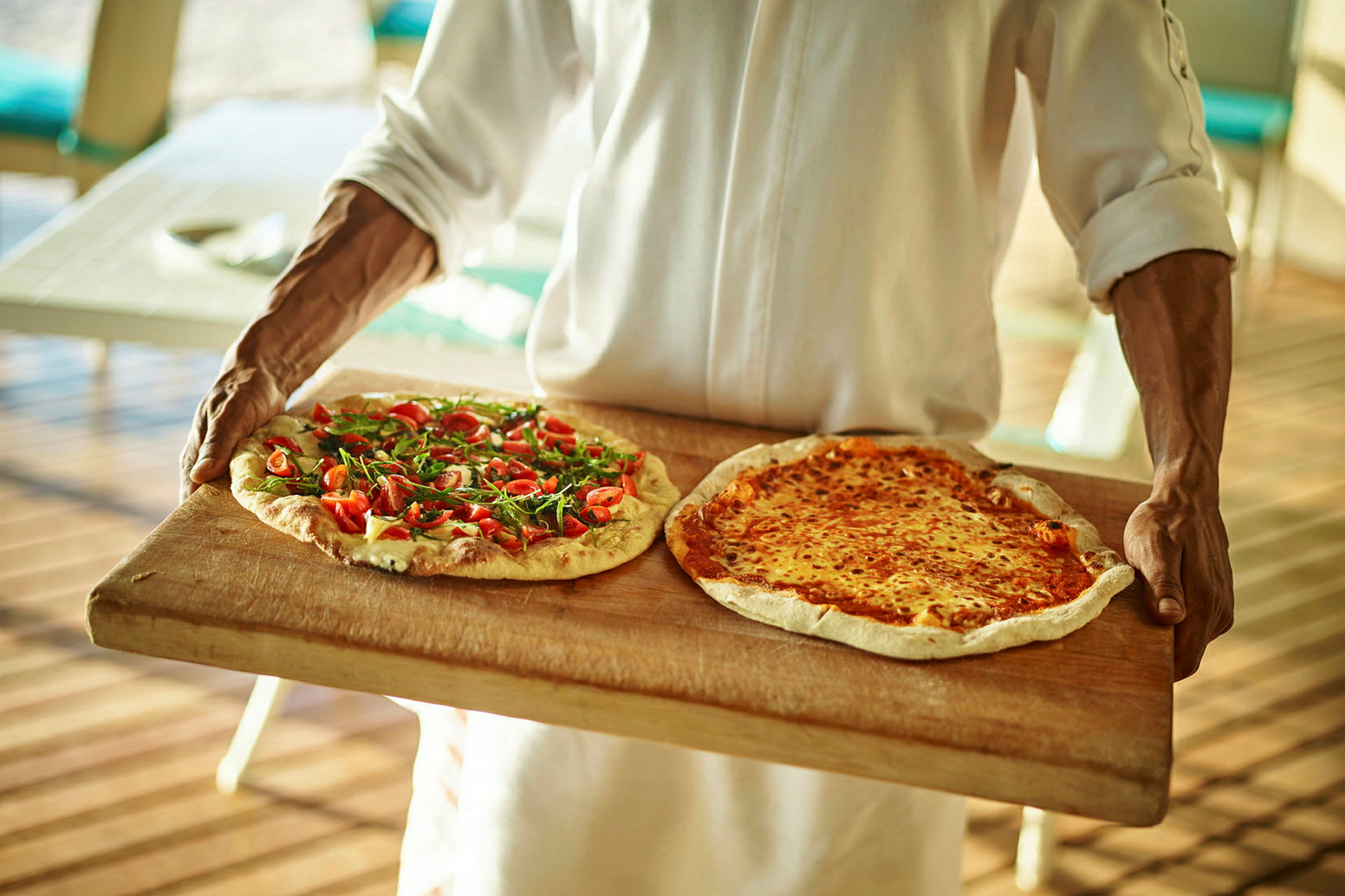 Henkilö kantaa kahta pizzaa, joista toisessa on tuoreita tomaatteja ja rucolaa ja toisessa sulatettua juustoa, puualustalla. (Tekoälyn luoma teksti)