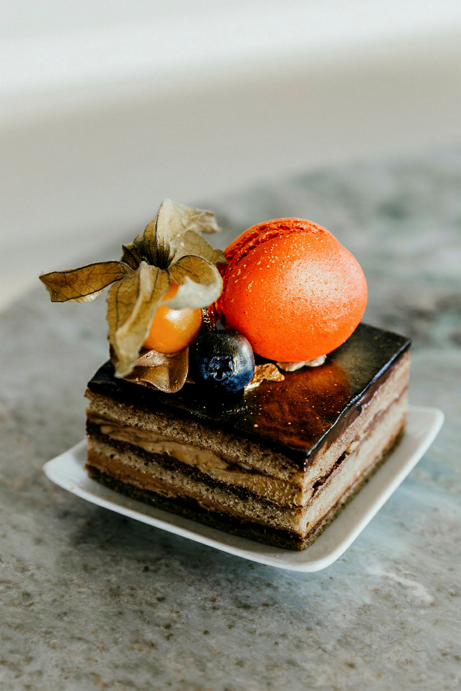 Lähikuva elegantista jälkiruoasta, jossa on suklaakuorrutus, kerroksia kermaa, koristeltuna oranssilla macaronilla, yksi karviainen ja mustikka. (Tekoälyn luoma teksti)