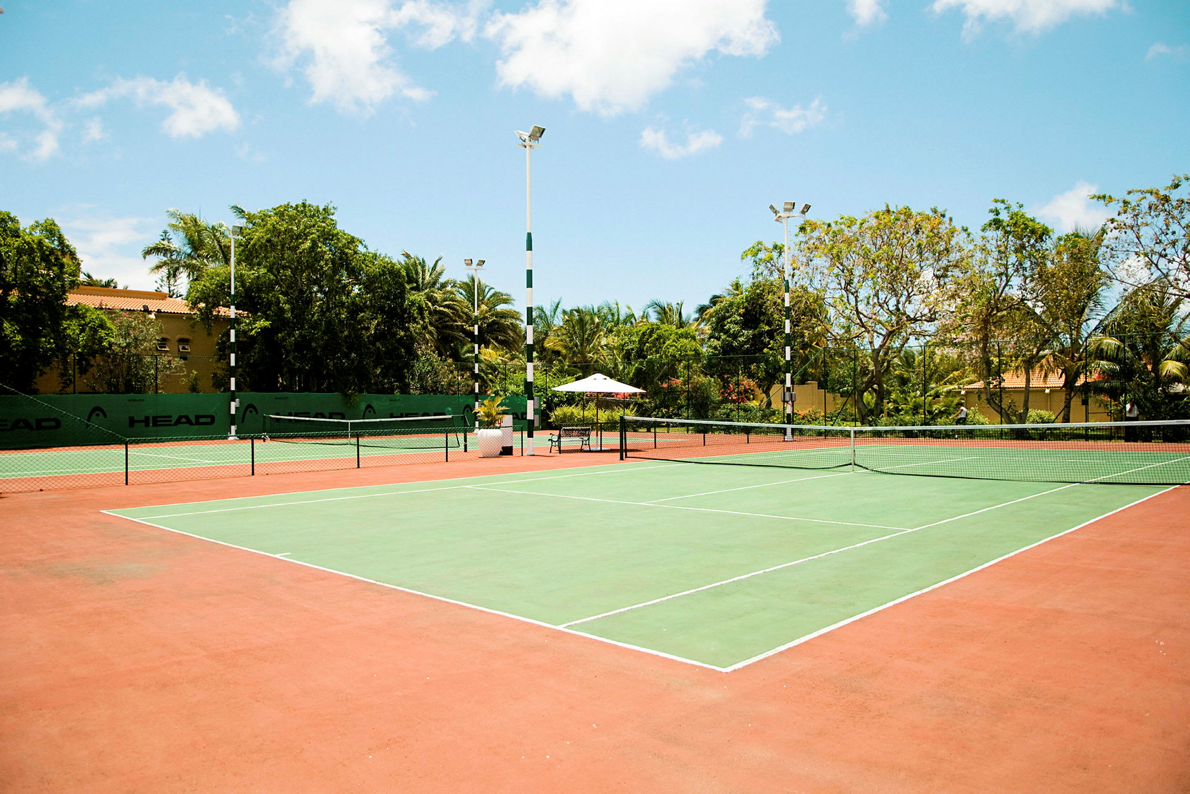 Tenniskenttä, jossa vihreä pelialue ja punaiset ympäröivät alueet, ympärillä puita ja rakennuksia. (Tekoälyn luoma teksti)