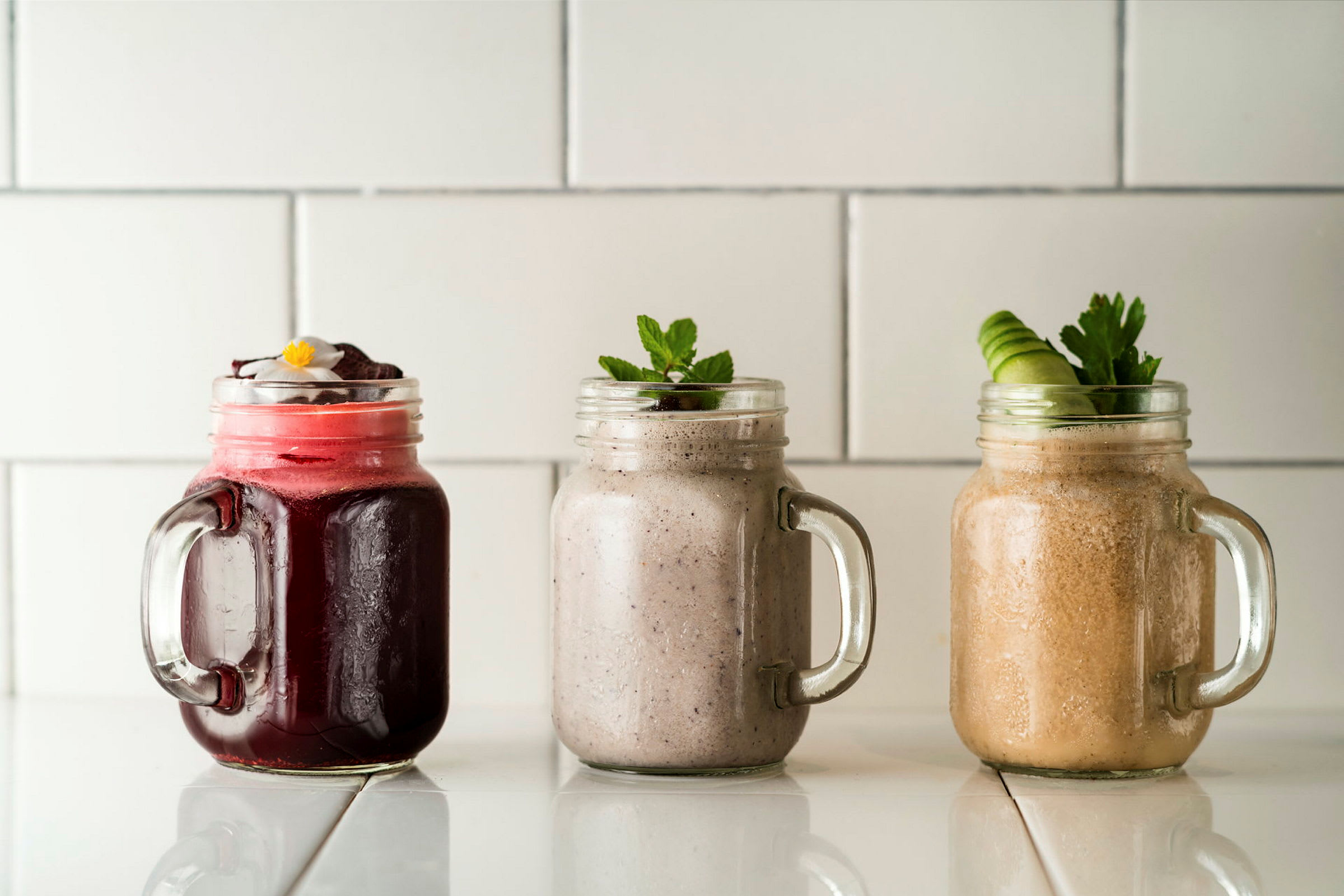 Kolme erilaista smoothiea lasipurkeissa valkoisella pinnalla. (Tekoälyn luoma teksti)