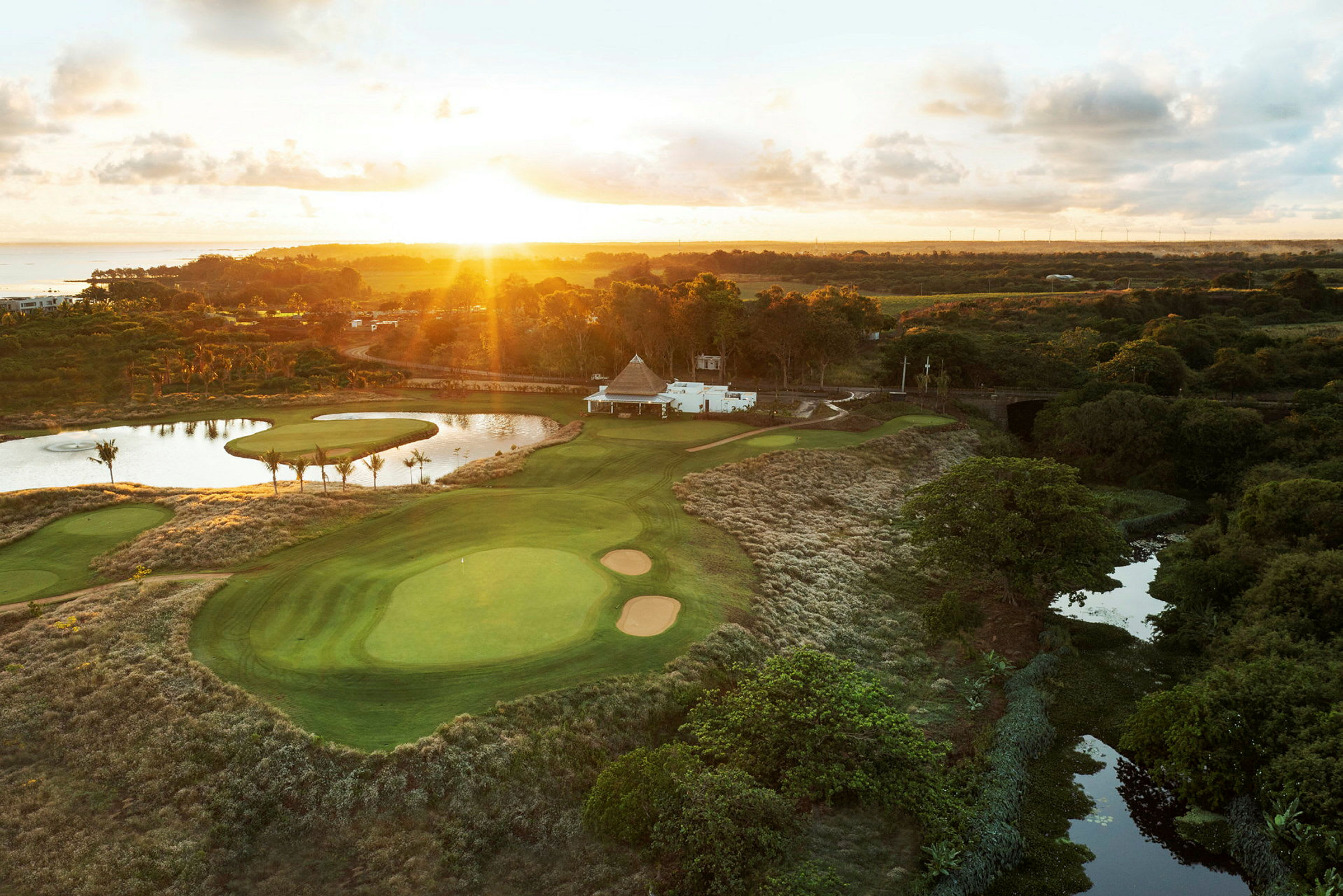 Golfkenttä, jolla vihreitä väyliä, bunkkereita ja lampi, ympärillä kasvillisuutta auringonlaskun aikana. (Tekoälyn luoma teksti)
