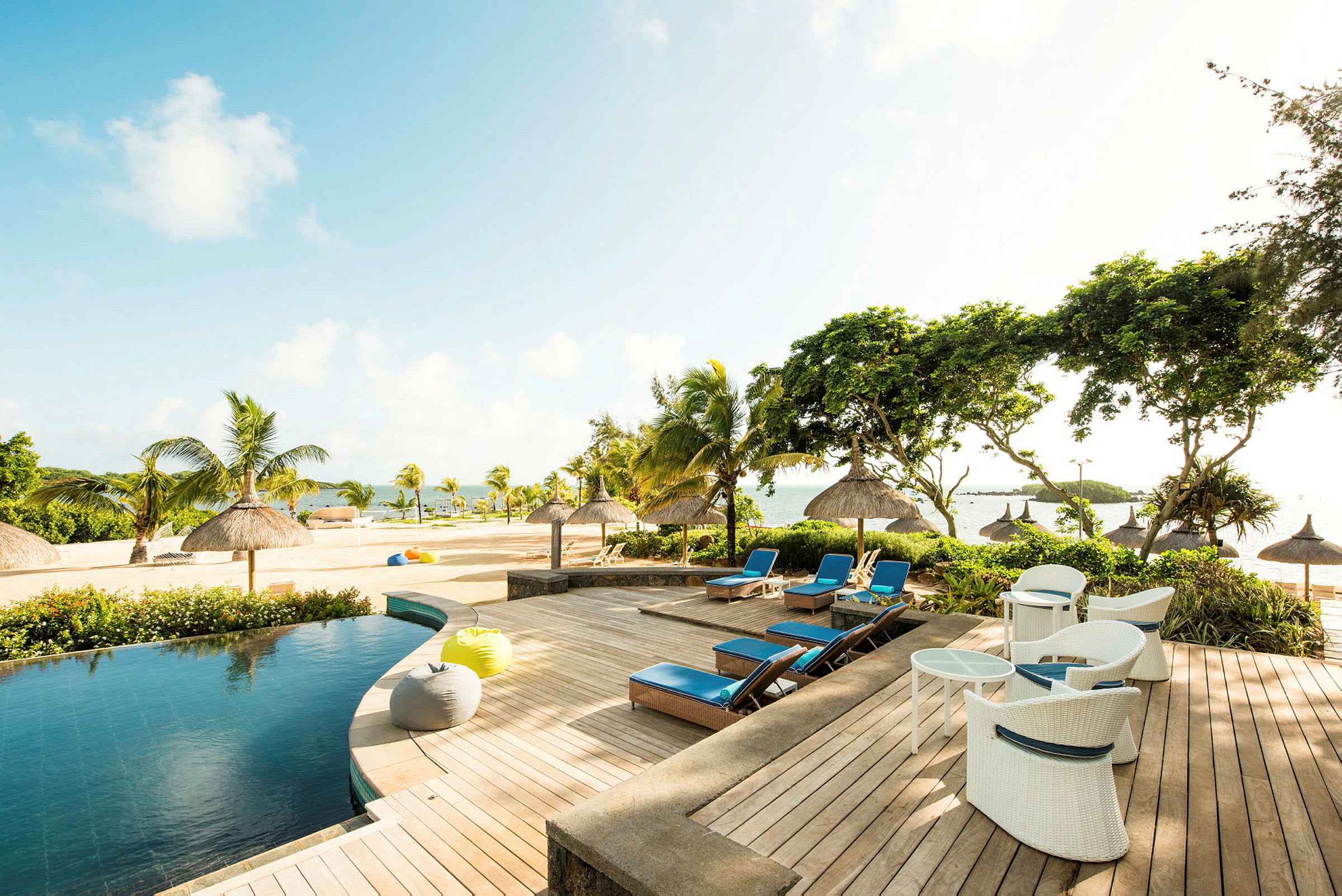 Allasalue meren rannalla, jossa on aurinkotuoleja, aurinkovarjoja ja vehreää ympäristöä. (Tekoälyn luoma teksti)