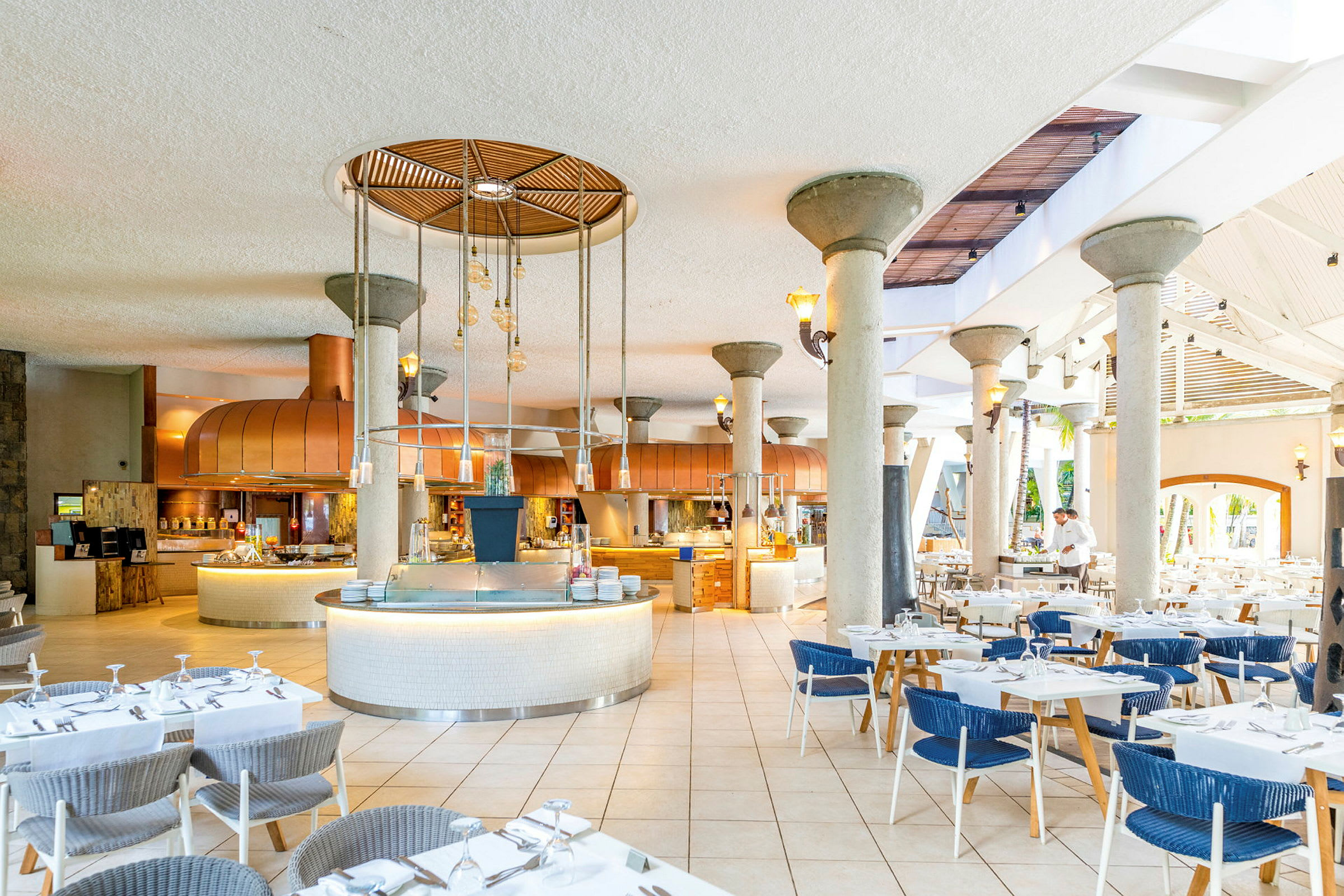 Ravintola, jossa on pöytiä ja tuoleja, keskellä sijaitseva tarjoilualue ja kattokoristeita. (Tekoälyn luoma teksti)
