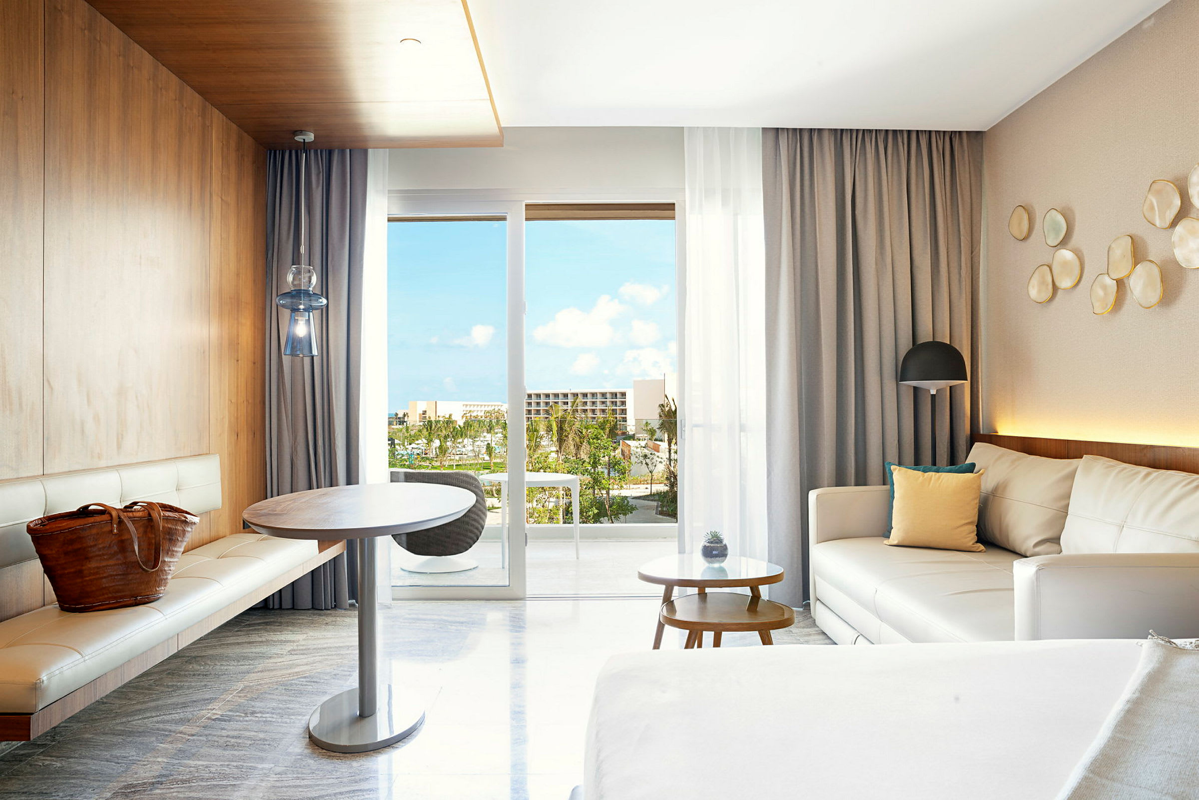 Moderni hotellihuone, jossa valkoiset huonekalut ja puurakenteet. Panoraamaikkunoista parvekkeelta näkyy trooppisia kasveja ja sininen taivas. (Tekoälyn luoma teksti)