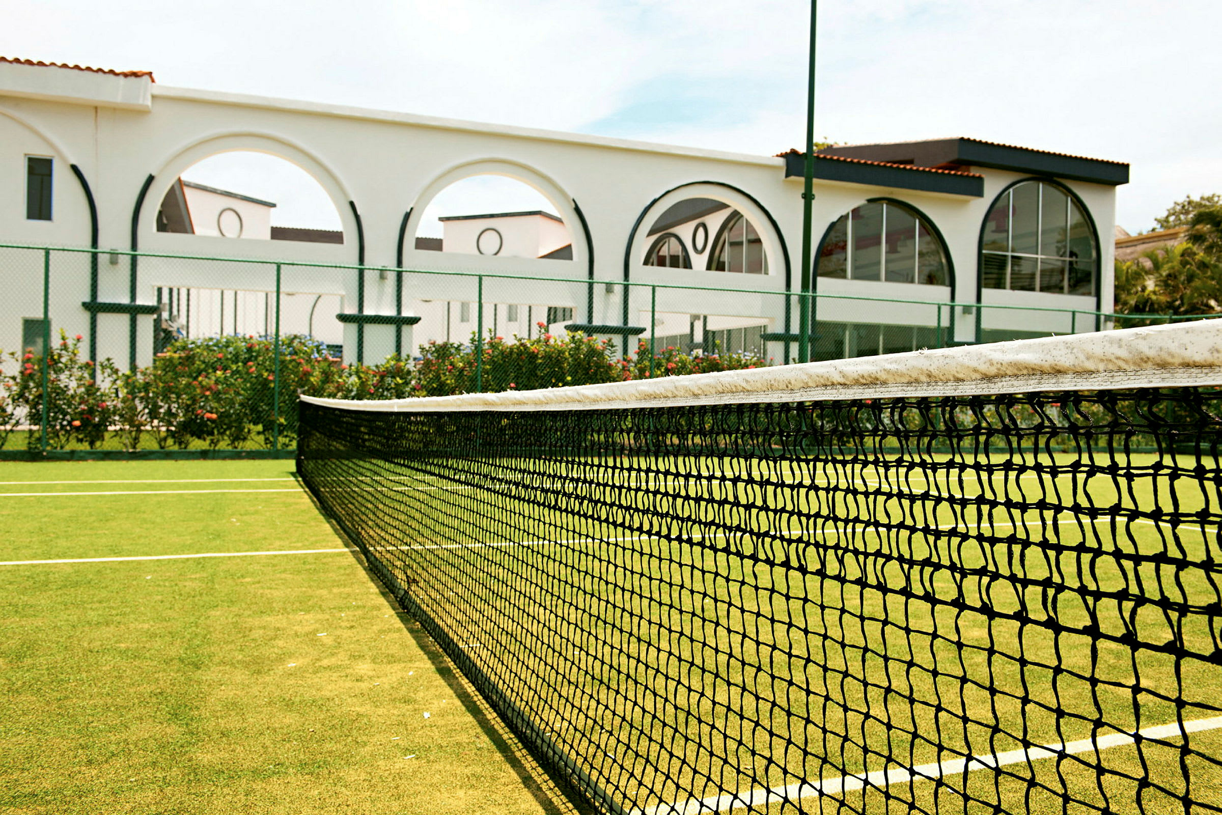 Tenniskenttä, jonka etualalla on verkko. Verkkojen takana näkyy valkoinen rakennus, jossa on kaaria ja lasia. (Tekoälyn luoma teksti)