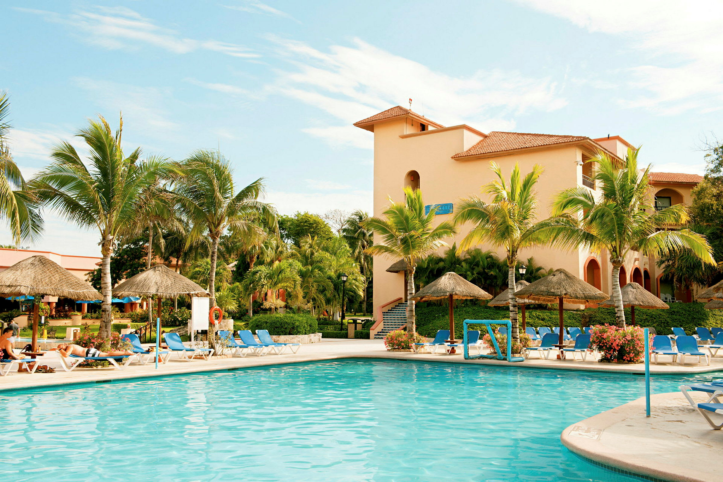 Suuri uima-allas, jota ympäröivät aurinkotuolit, palmut ja aurinkovarjot. Allasalueen takana on rakennus terrakotan värisillä seinillä. (Tekoälyn luoma teksti)