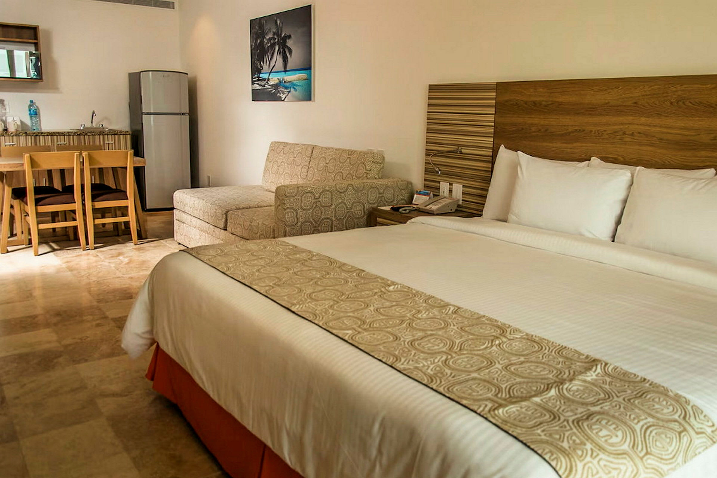 Hotellihuone, jossa on suuri sänky, istuimia, pöytä ja pieni keittiö jääkaapilla. (Tekoälyn luoma teksti)