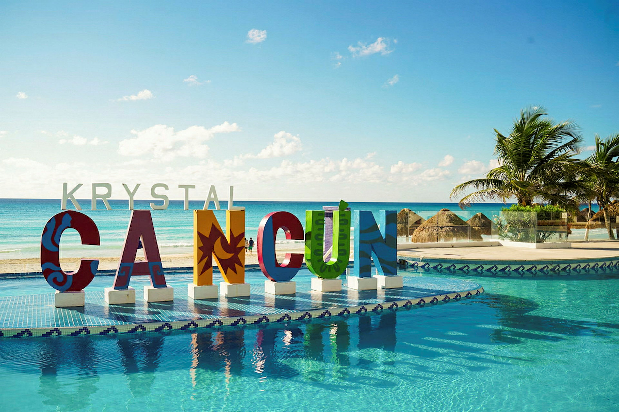 Värikkäitä kirjaimia, jotka muodostavat sanan 'CANCUN' altaan äärellä merinäköalalla ja palmuja. (Tekoälyn luoma teksti)
