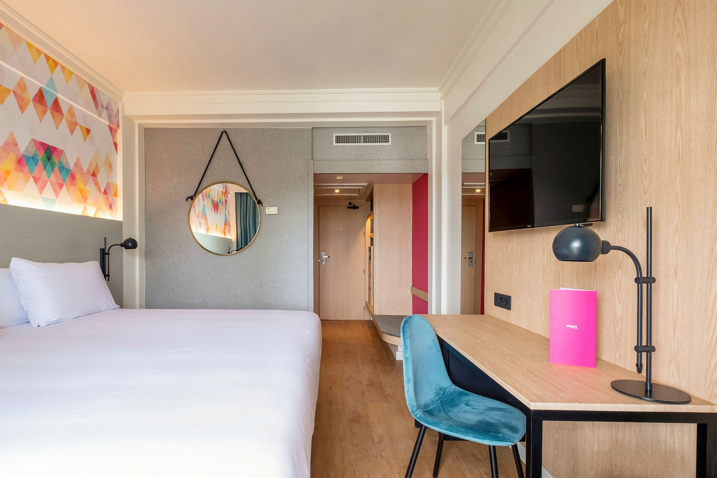 Hotellihuone, jossa on sänky, työpöytä tuolin ja lampun kanssa sekä seinälle asennettu televisio. (Tekoälyn luoma teksti)