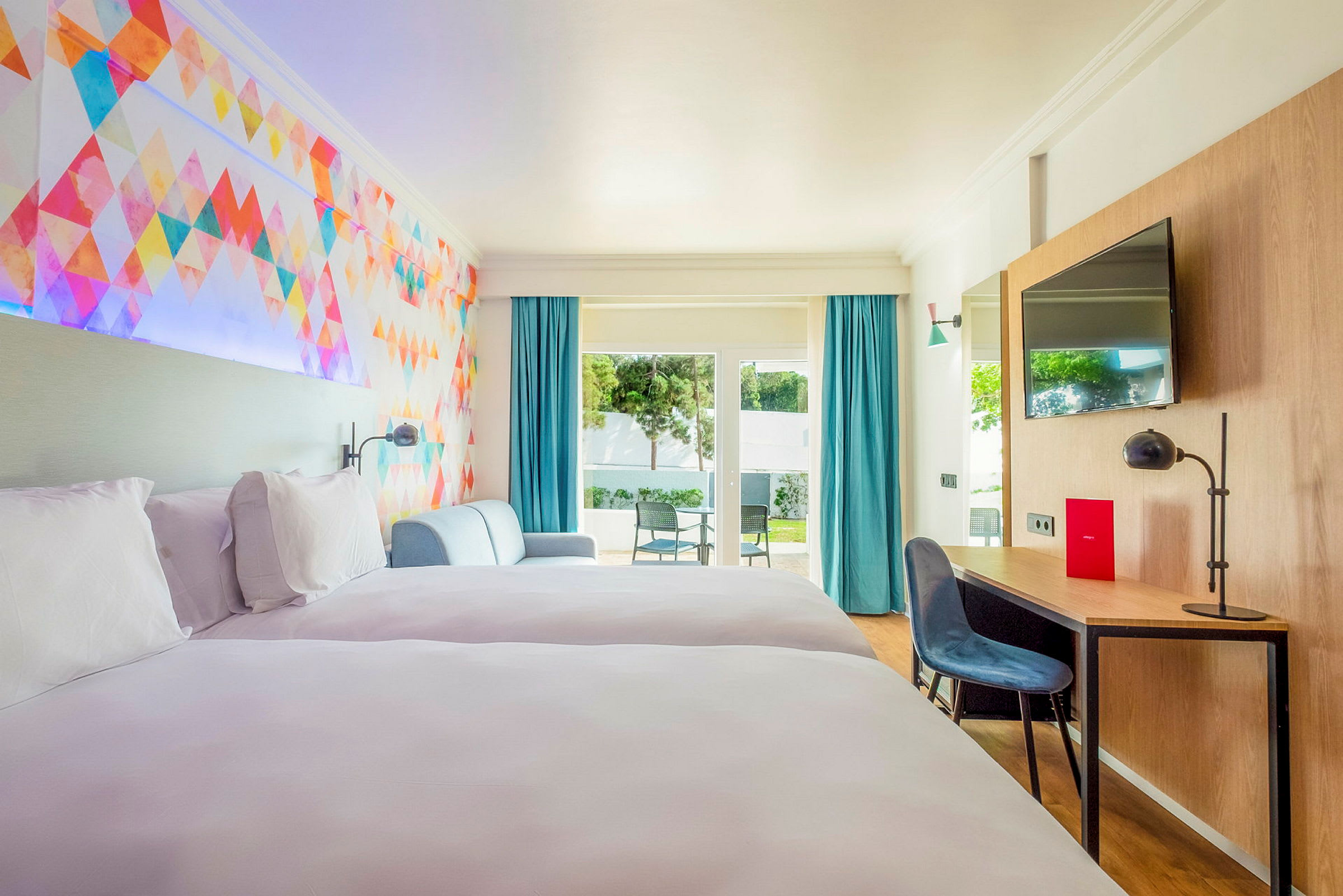 Hotellihuone, jossa kaksi sänkyä, värikäs seinäkoriste, oleskelutila, työpöytä ja pääsy terassille, jossa on tuoleja. (Tekoälyn luoma teksti)