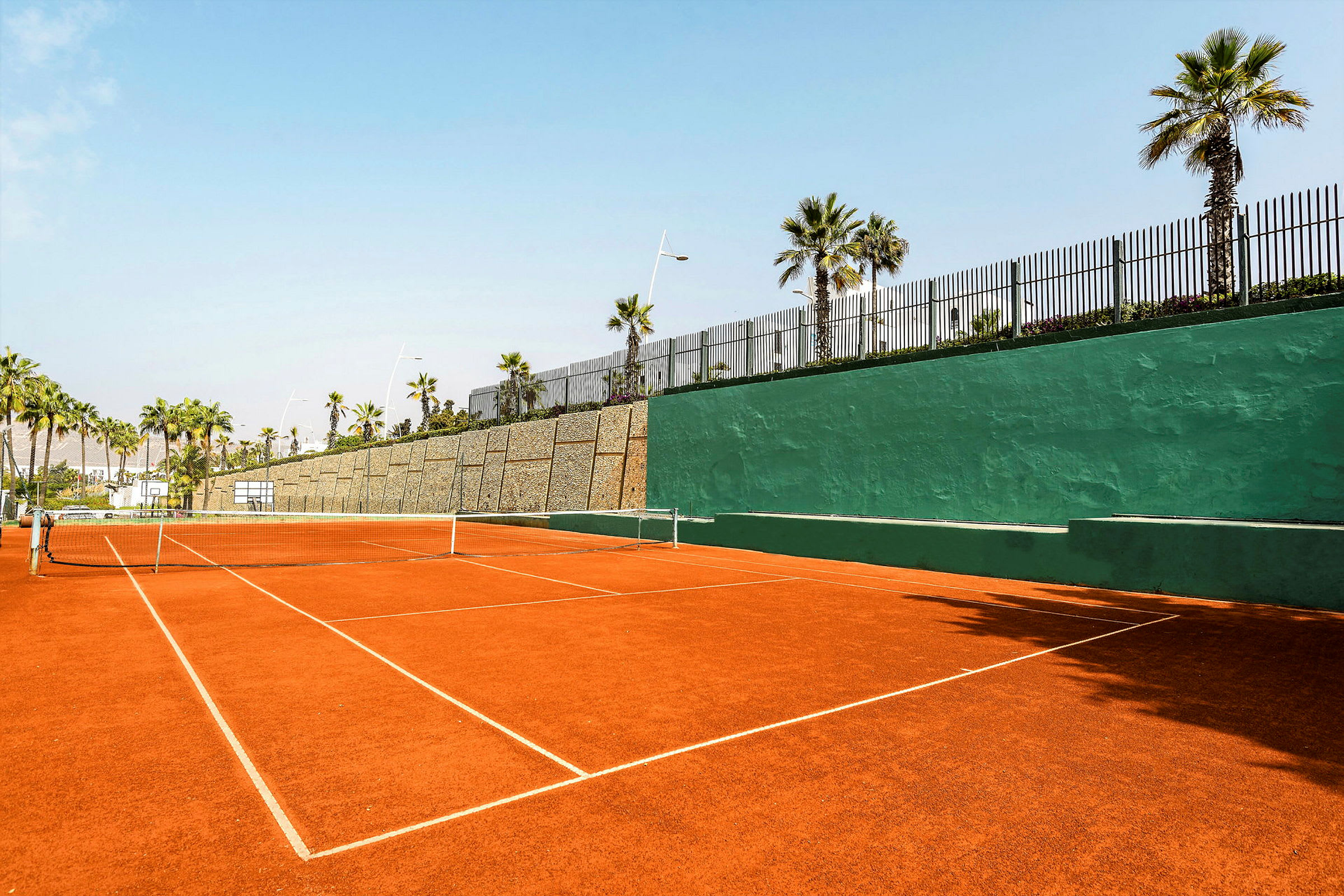 Tenniskenttä, jossa on punainen sora ja vihreä seinä taustalla, ympärillä palmuja. (Tekoälyn luoma teksti)