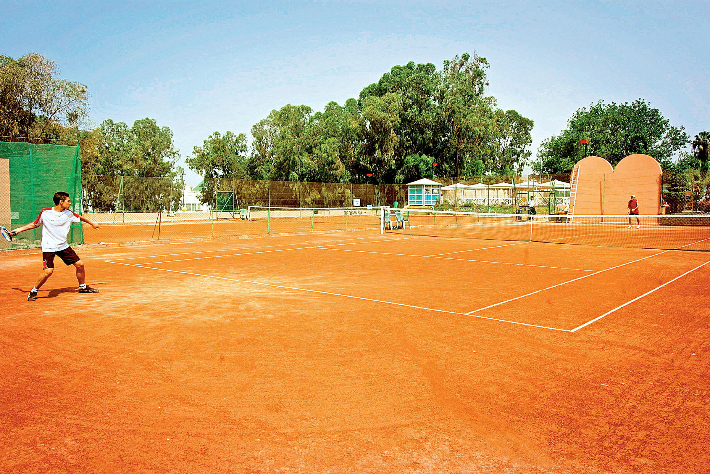 Ulkotenniskenttä, jossa kaksi henkilöä pelaa tennistä. (Tekoälyn luoma teksti)